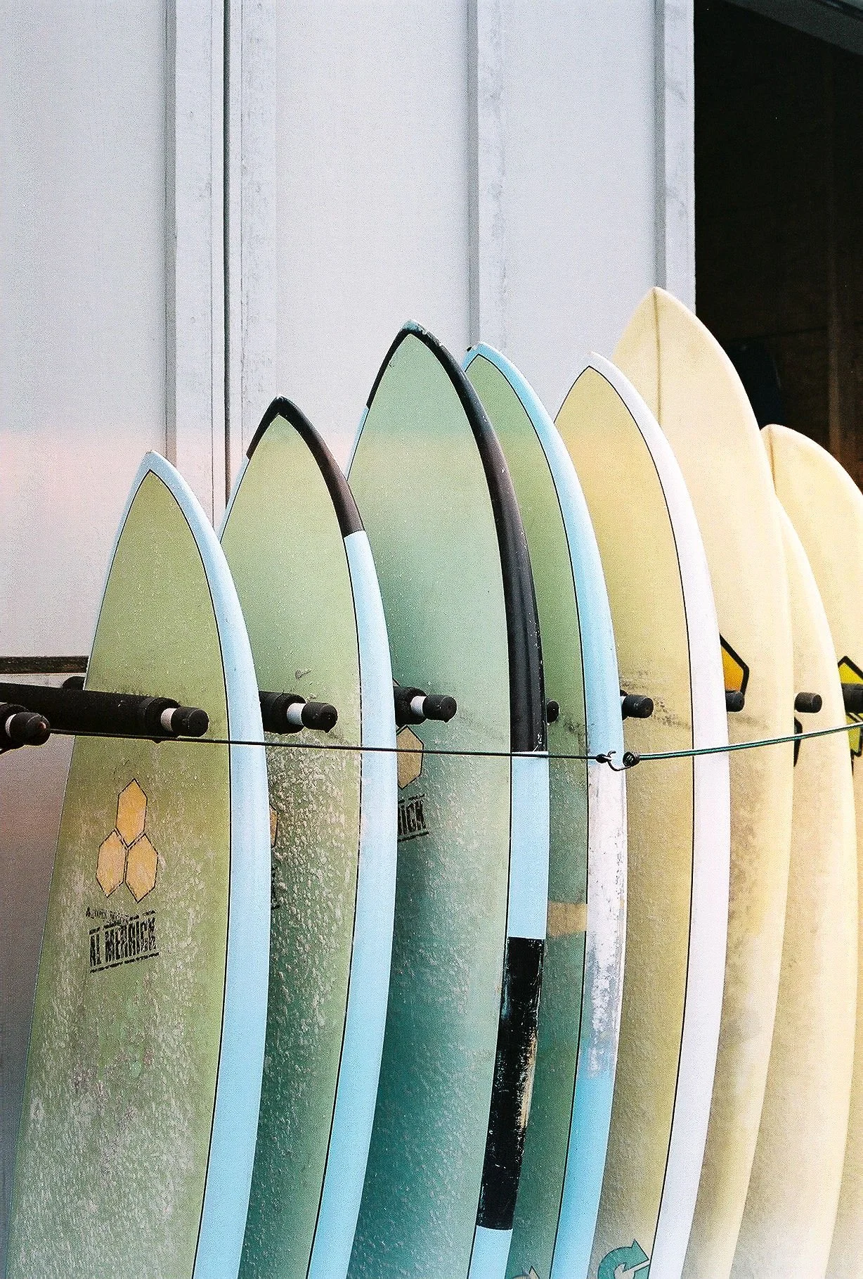 A row of colorful surfboards standing upright against a white wall, with some tires and a wire running across the boards.