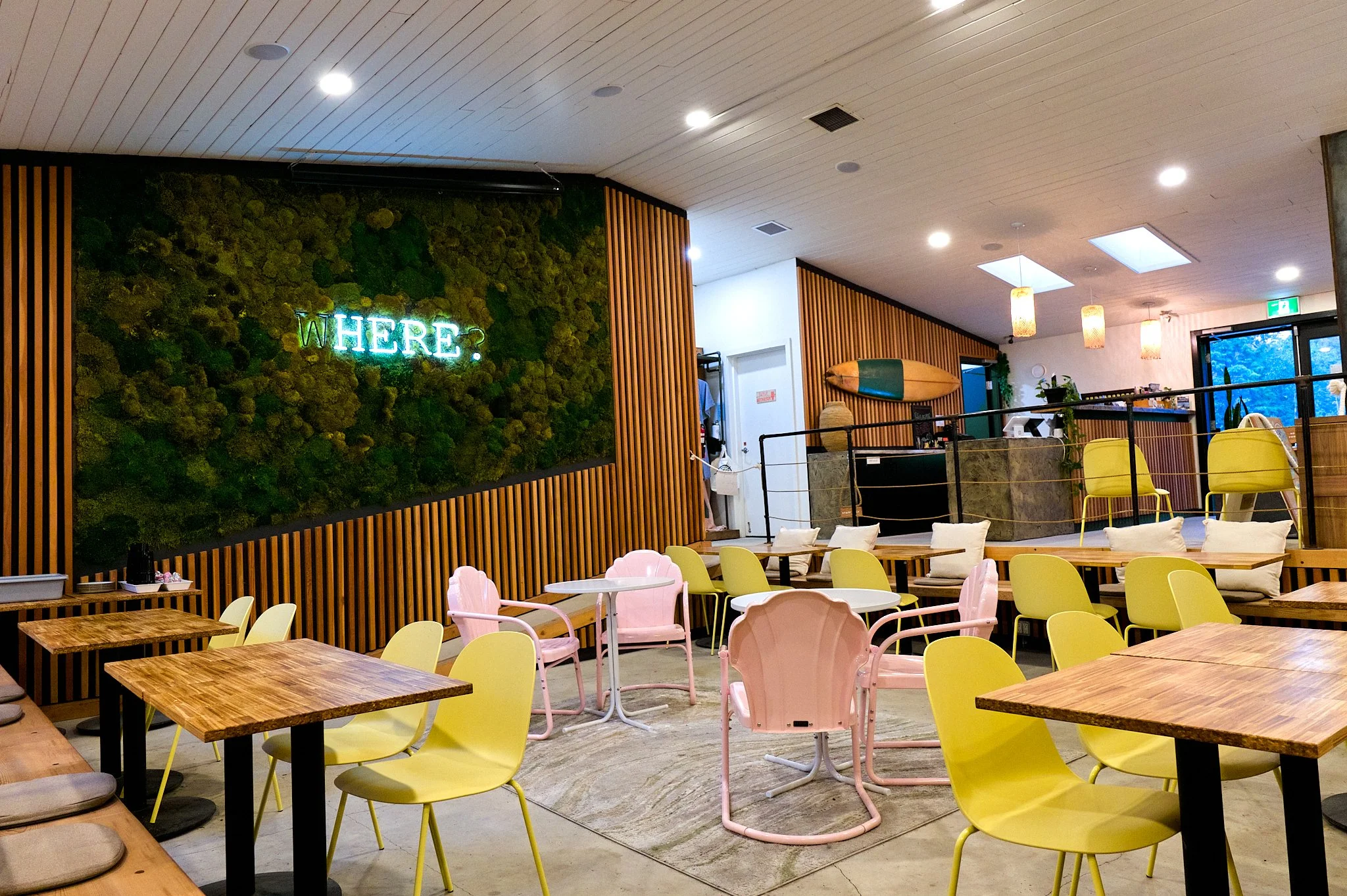 Interior of a cafe with wooden tables and colorful chairs, a moss wall with neon sign that says "WHERE?", surfboard decoration on the wall, and a counter area in the background.