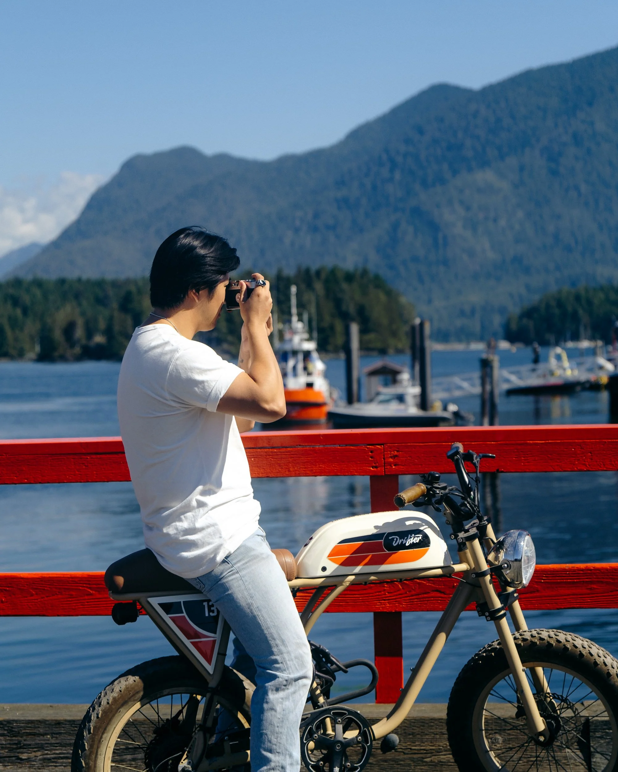Person sitting on a vintage-style electric bicycle by a river, taking a photo with a camera, with mountains and boats in the background.