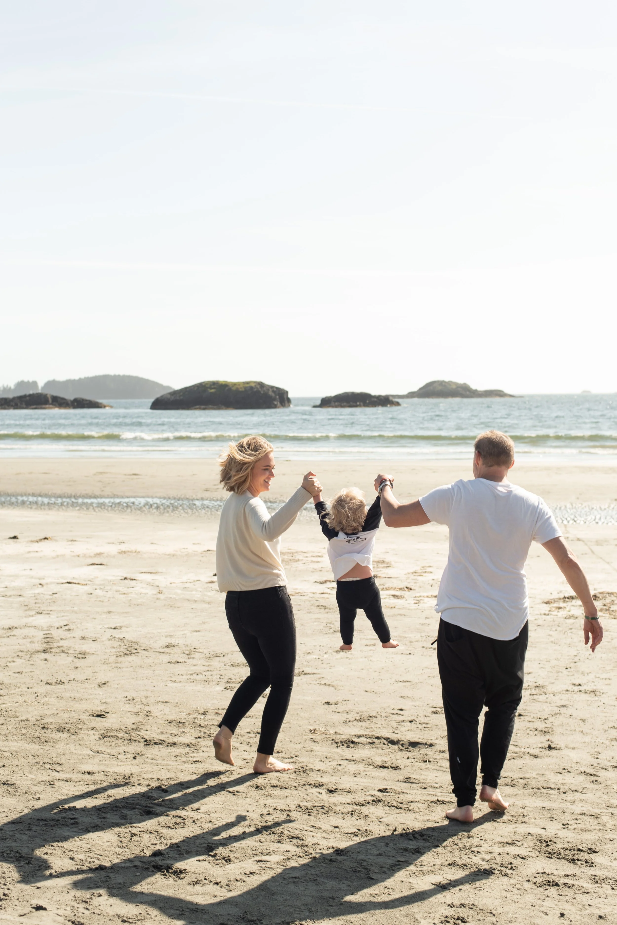 A family of three, two adults and a child, playing on a sandy beach during the day, with the adults holding the child's hands and swinging them in the air. The ocean and distant rocks are visible in the background.