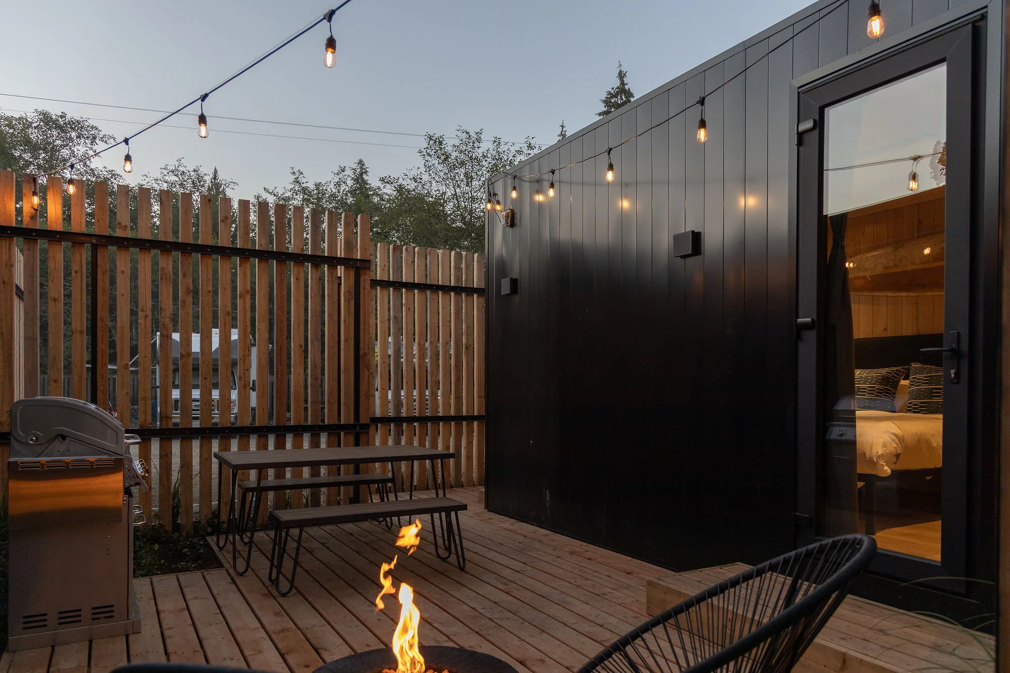 A cozy outdoor patio with string lights, a small fire pit, a grill, a picnic table, and a black modern guest house with large windows, surrounded by a wooden fence and trees.