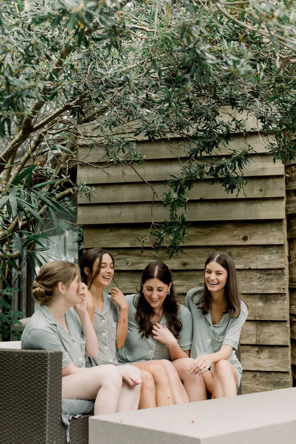 Four women sitting on a couch outdoors, wearing pajamas, laughing and talking, with a wooden fence and leafy tree in the background.