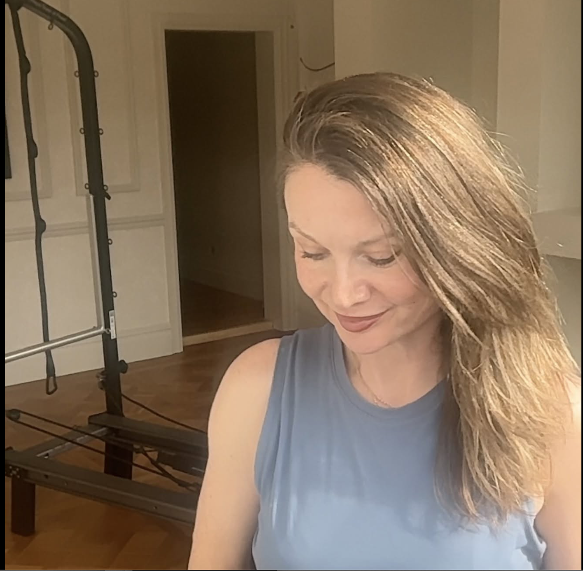 A woman with long, blonde hair looking down and smiling, wearing a blue sleeveless top, standing in a room with a door and a shelving unit in the background.