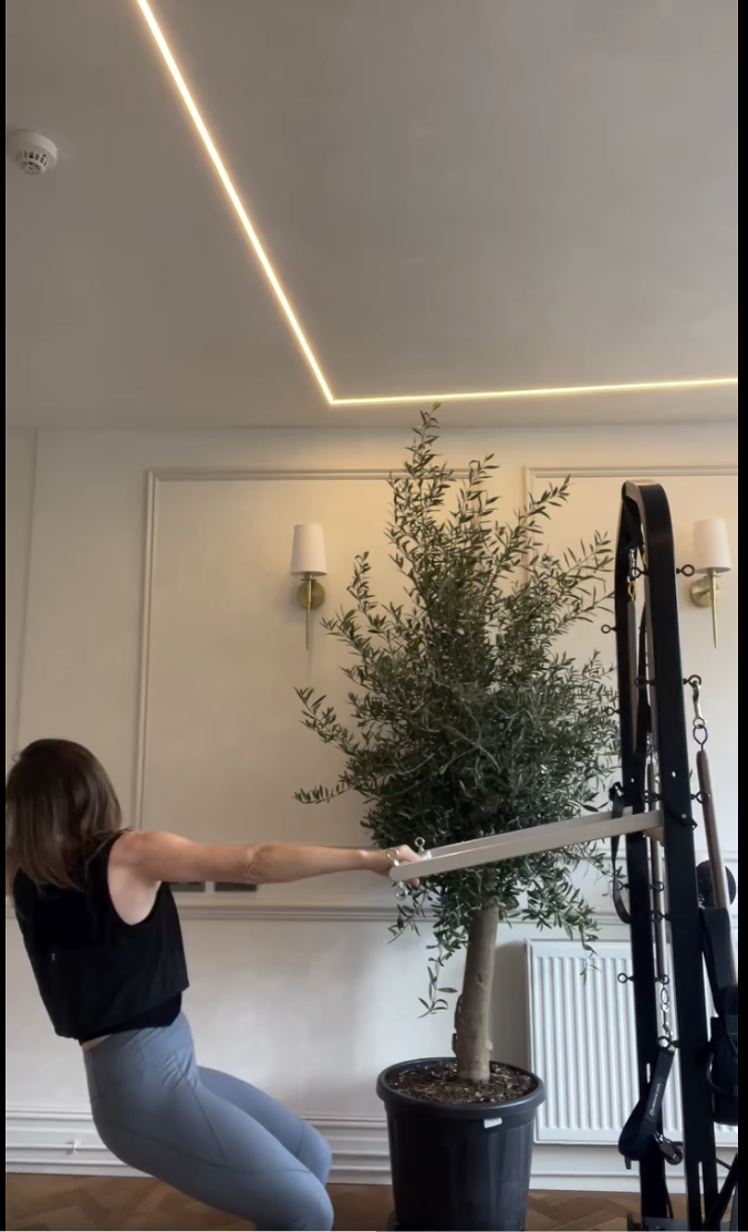 Woman in workout clothes pulling on a resistance band attached to a large potted tree indoors, with decorative wall lights and a ceiling with LED strip lighting.