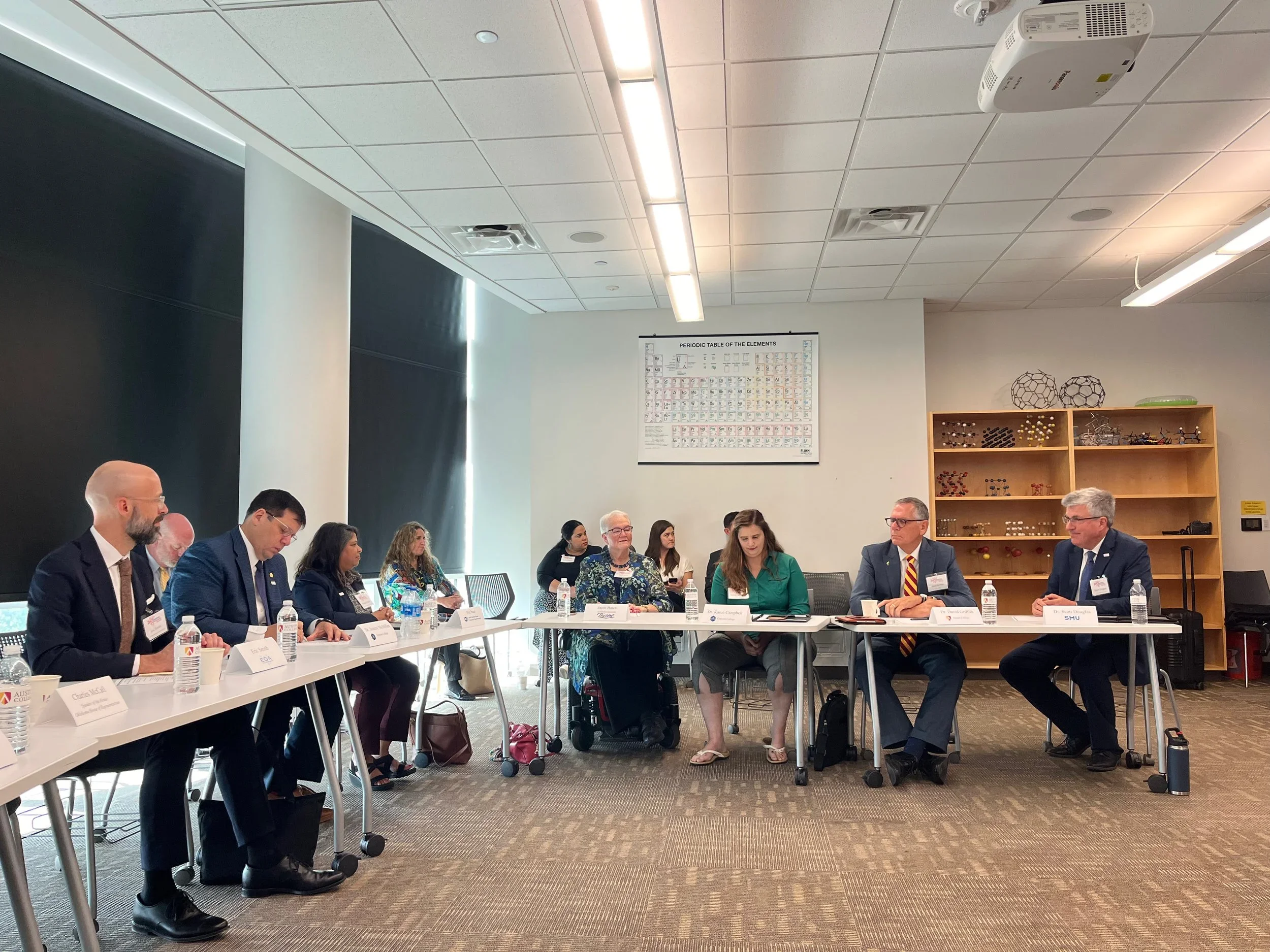 A group of diverse adults seated around a conference table in a meeting room, engaged in discussion. Water bottles and name tags are in front of them, with a poster of the periodic table on the wall and shelves with decorative objects in the background.