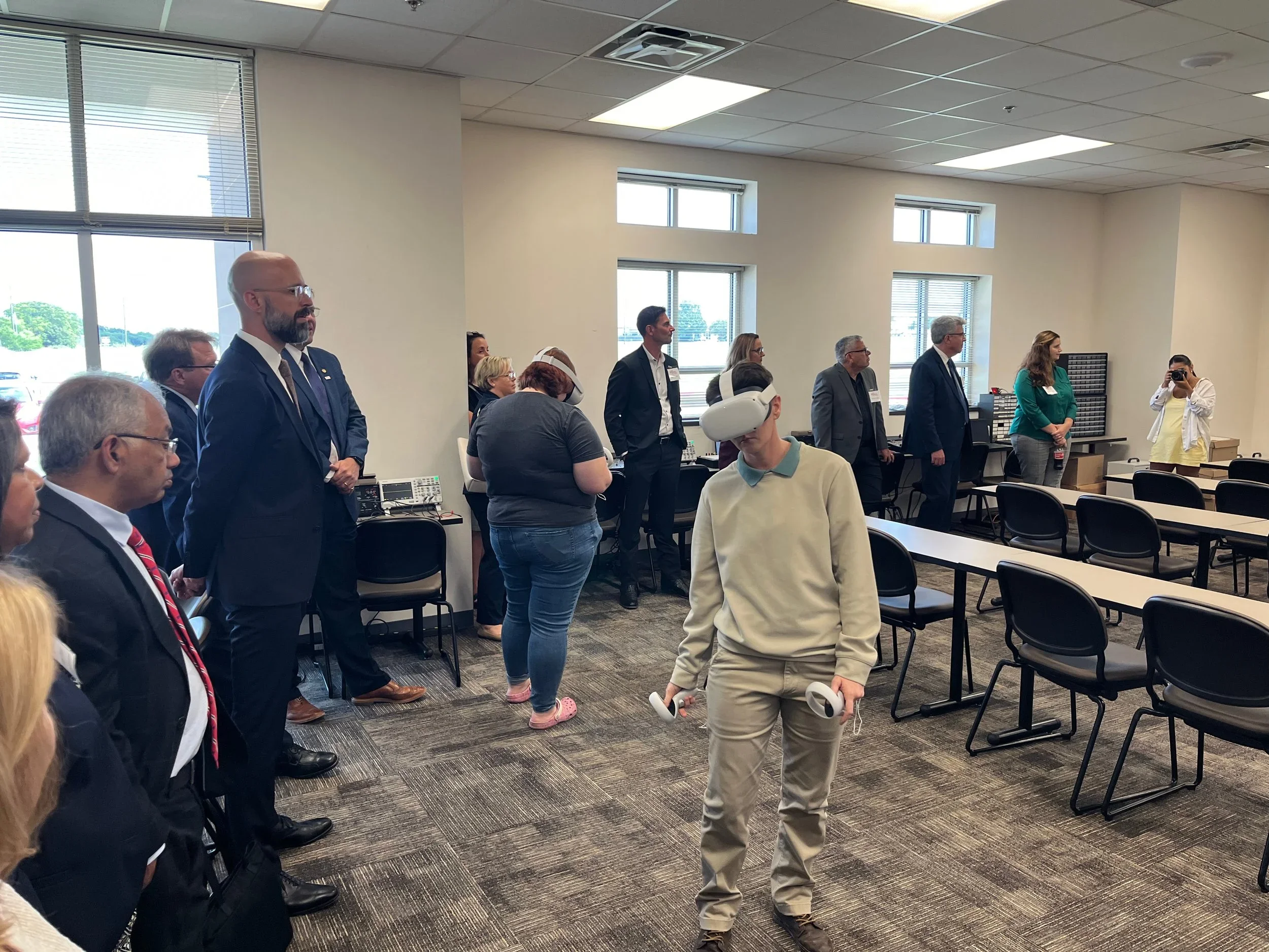 A young man wearing a VR headset and holding a VR controller stands in the foreground of a conference room filled with professionally dressed people, some standing and some seated. The room has large windows, black chairs, and a gray carpet.
