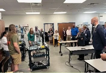 Group of people in a classroom or training room, standing and sitting around tables, with medical equipment visible in the background.