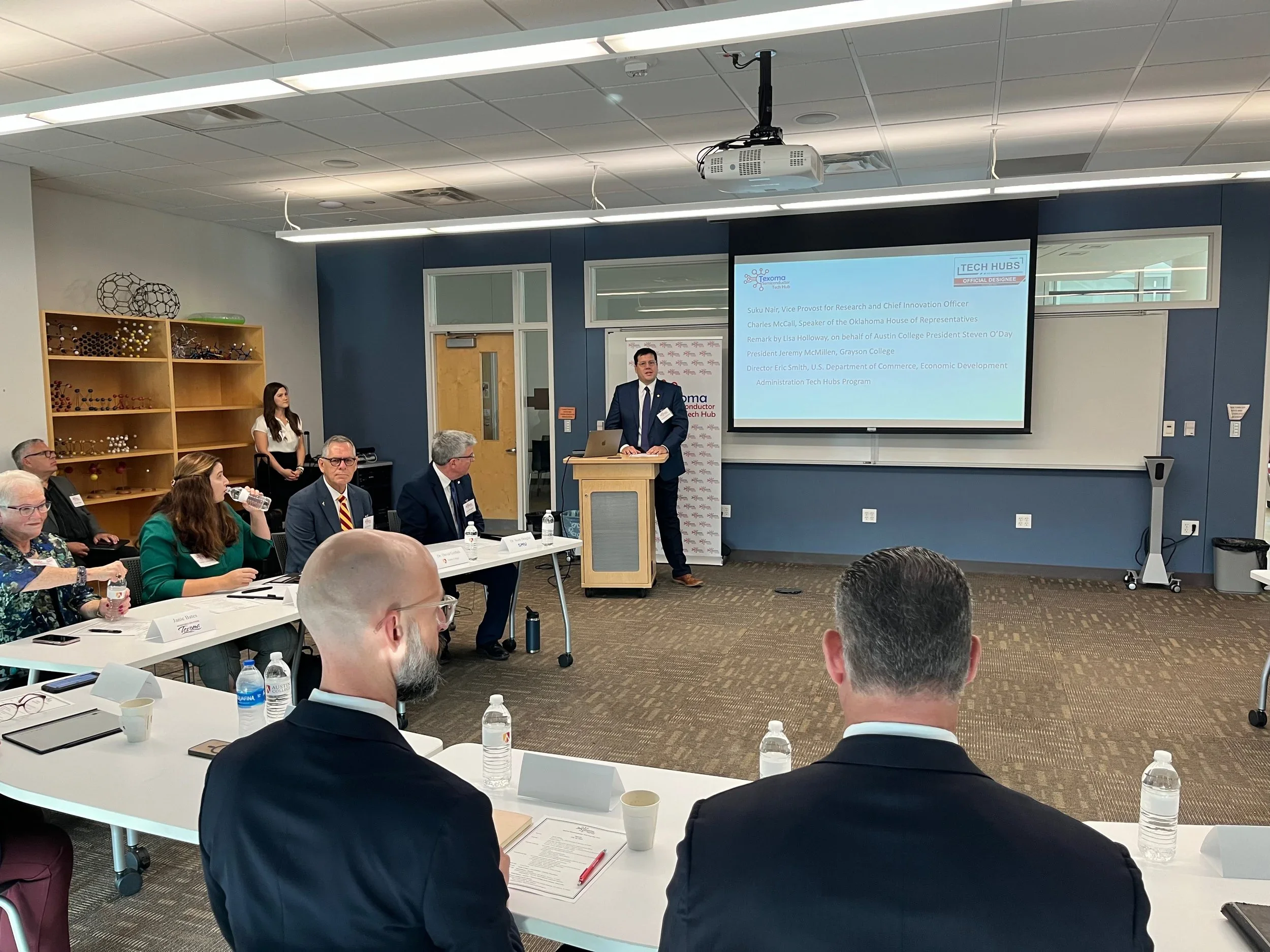 A man in a suit stands at a podium giving a presentation to an audience seated at tables in a conference room. A large screen behind him displays the agenda, with a few people looking towards him. Attendees include men and women, some with notebooks and water bottles, and a bookshelf with molecular models is visible on the left.