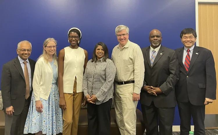 Group of seven diverse professionals standing together in front of a blue wall, smiling for the camera.