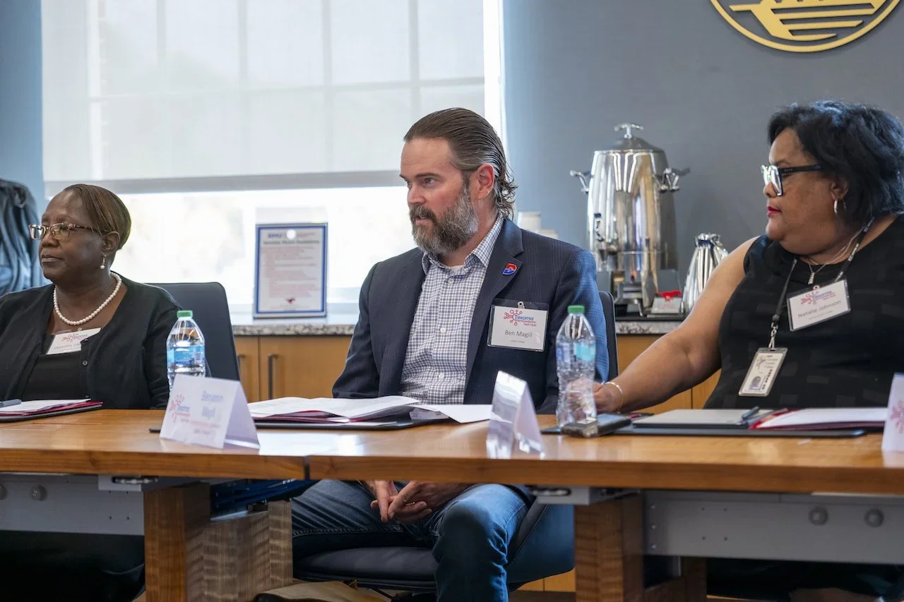 Three people sitting at a conference table during a meeting, with water bottles, name tags, and documents in front of them. There is a window in the background and a coffee station to the right.