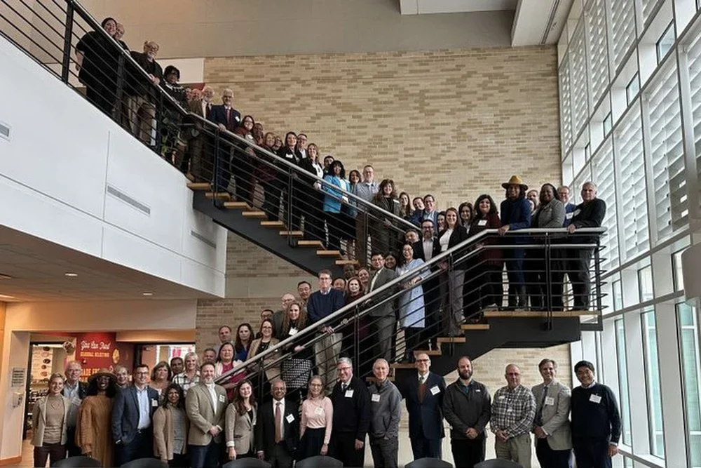 A large group of people, consisting of men and women, are gathered on a staircase inside a building with large windows and brick walls, posing for a group photo.