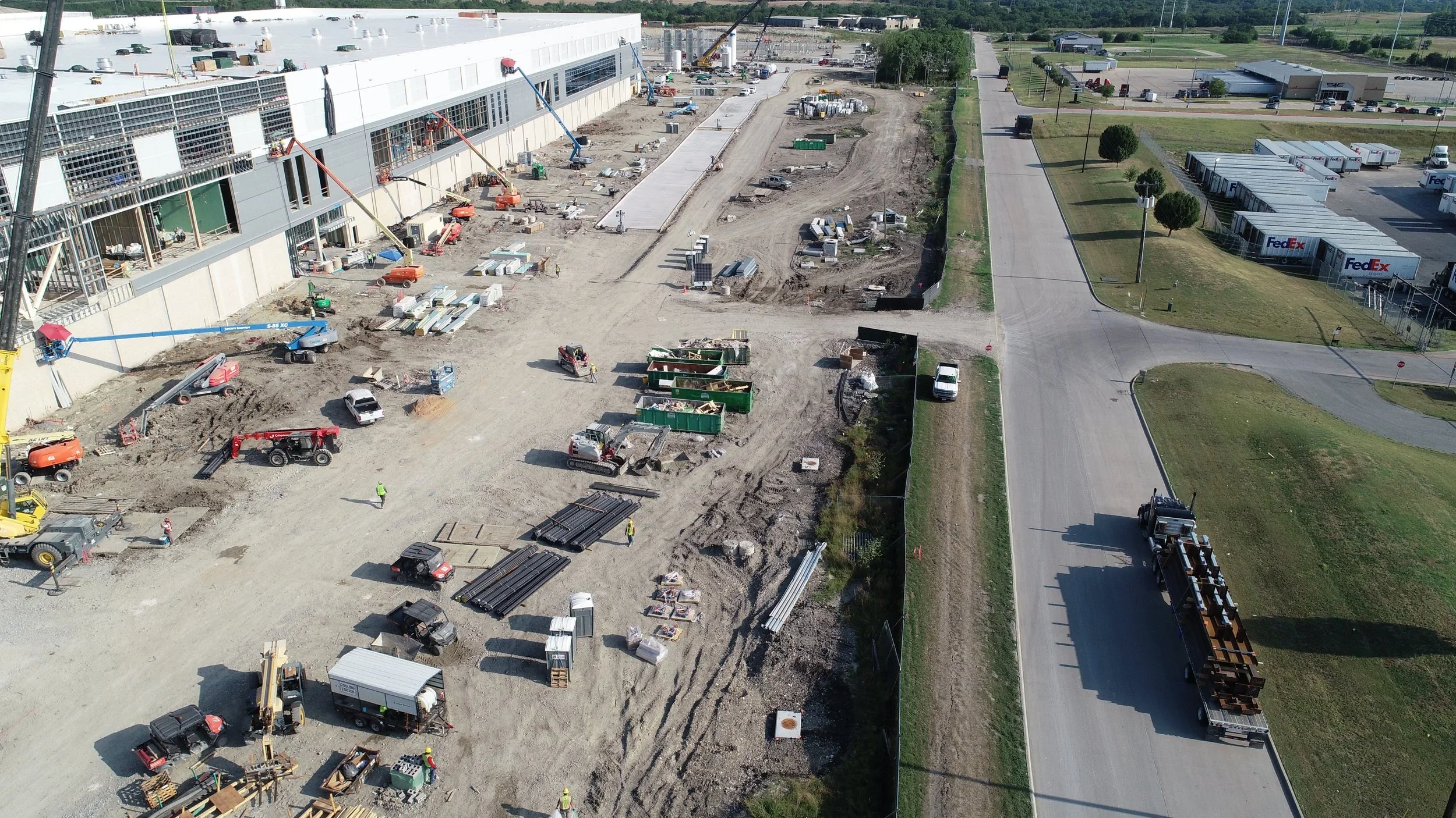Aerial view of a large construction site next to a commercial building with cranes, dump trailers, construction vehicles, and workers, with a paved road and FedEx distribution center in the background.