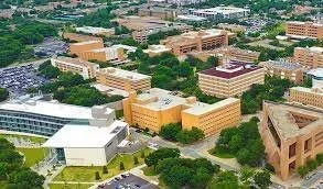 Aerial view of a university campus with multiple beige and brown brick buildings surrounded by trees and parking lots.