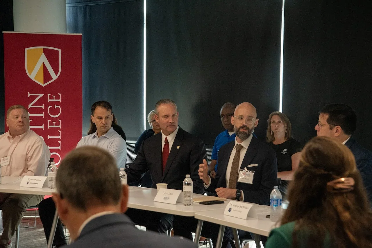 People participating in a panel discussion or meeting in a conference room, with water bottles and name tags on the table, and a red banner with a shield logo and the words 'Winning College' on the side.