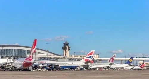 Multiple airplanes parked at an airport tarmac with a control tower and terminal in the background under blue sky.