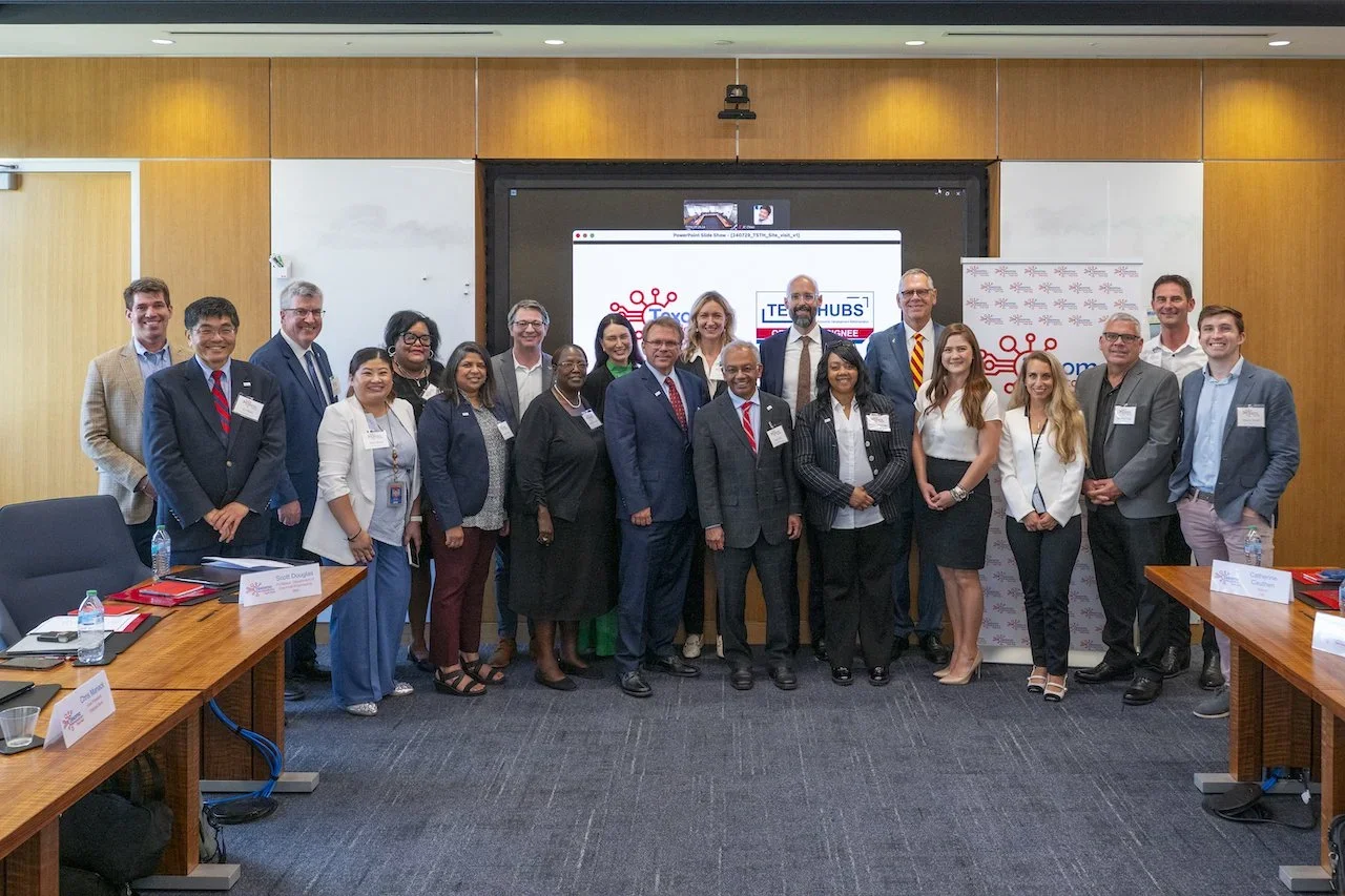 A group of diverse professionals posing for a photo in a conference room with a screen displaying the TEXHUBS logo in the background.