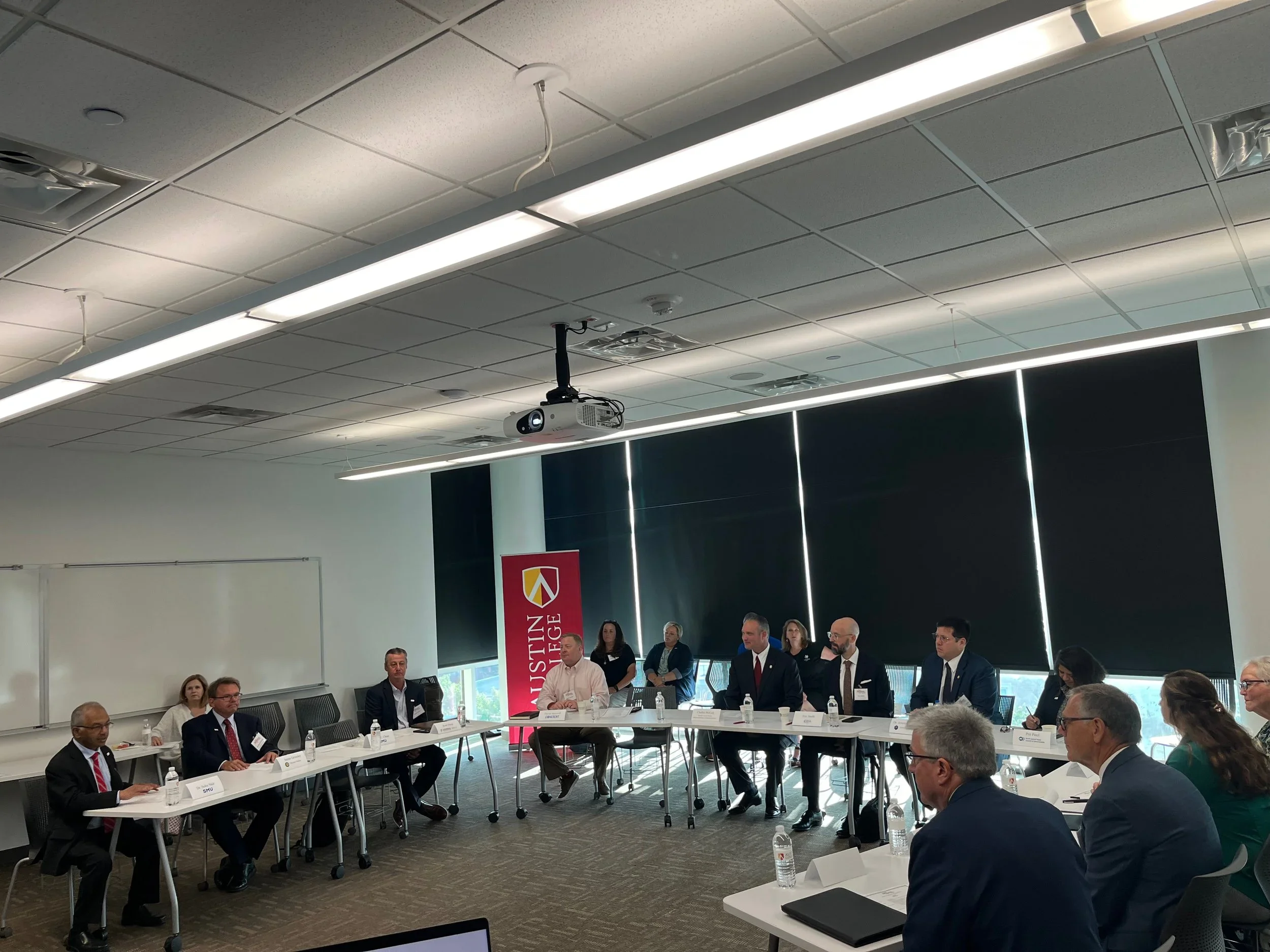 A group of people seated around a U-shaped conference table in a modern meeting room at Austin College, with a banner, water bottles, and laptops visible.