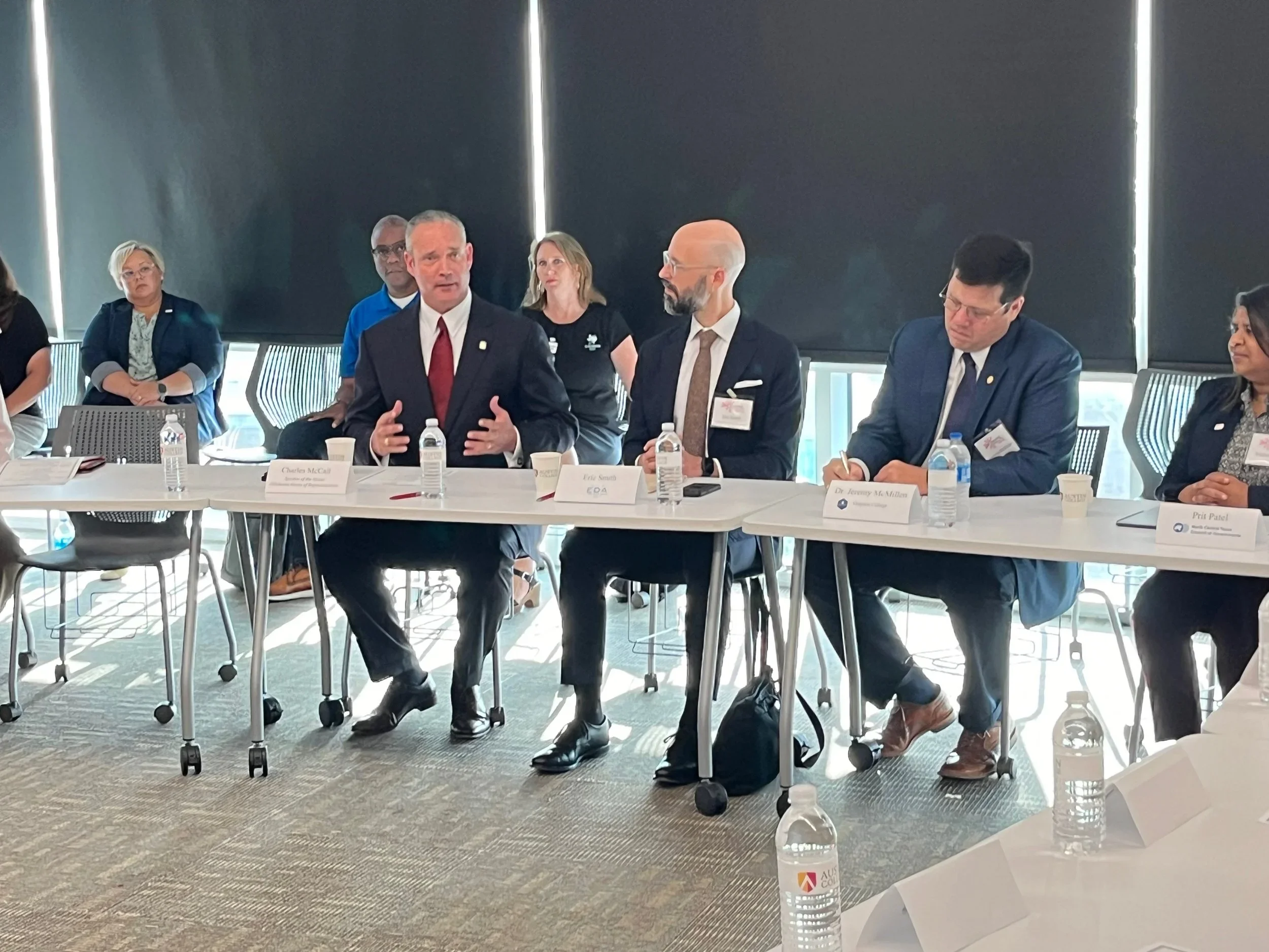 Business professionals seated at a long conference table in a meeting room with large window blinds behind them. One person is speaking, and others are listening or taking notes. Water bottles, notepads, and nameplates are on the table.