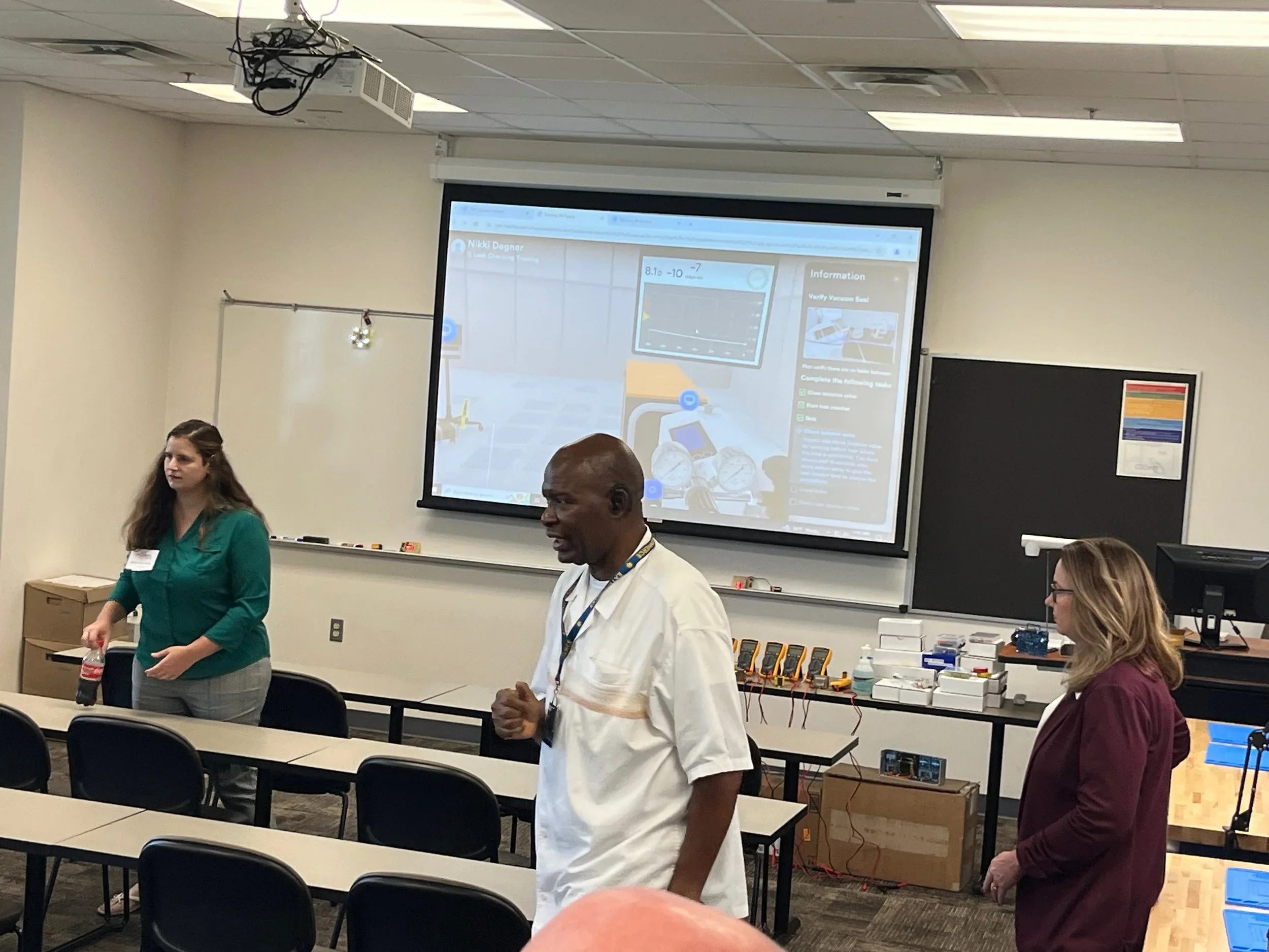 A classroom with three women and one man, some tables and chairs, a large projection screen displaying a presentation, and laboratory equipment on a table at the back.