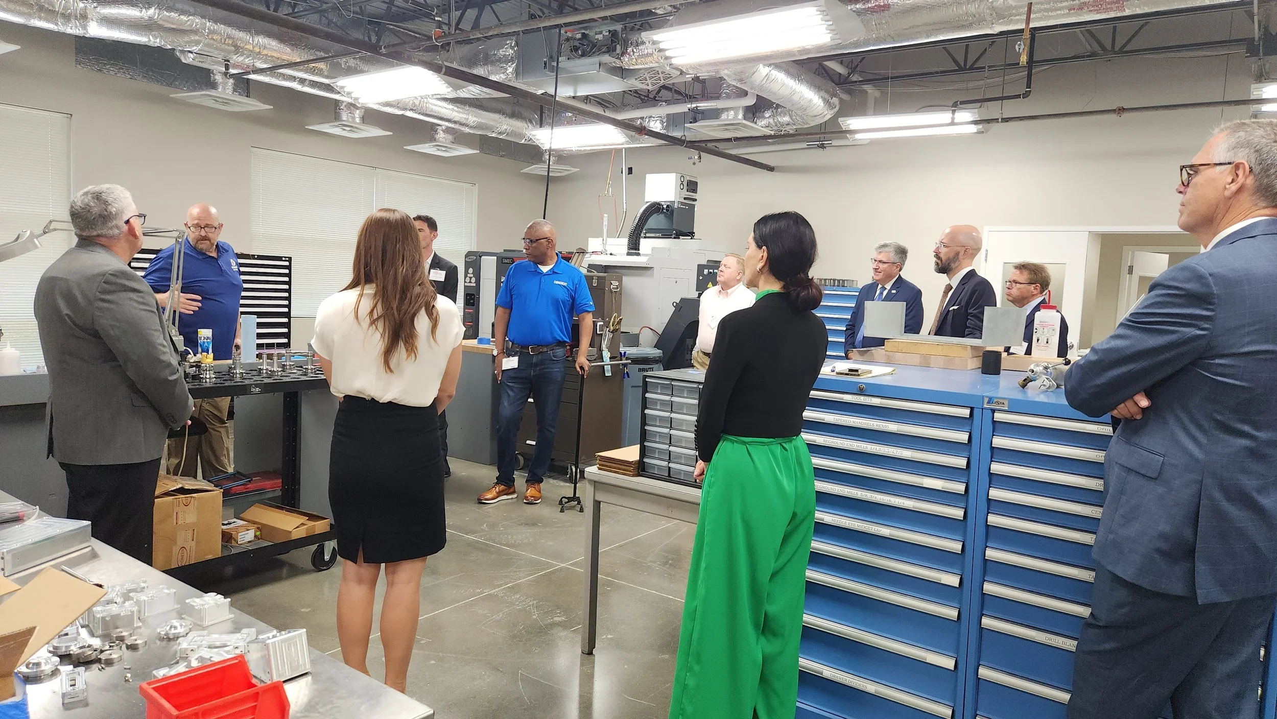 Group of people gathered in a manufacturing or industrial workshop, listening to a presentation or discussion. Tools and equipment are visible on workbenches and shelves.