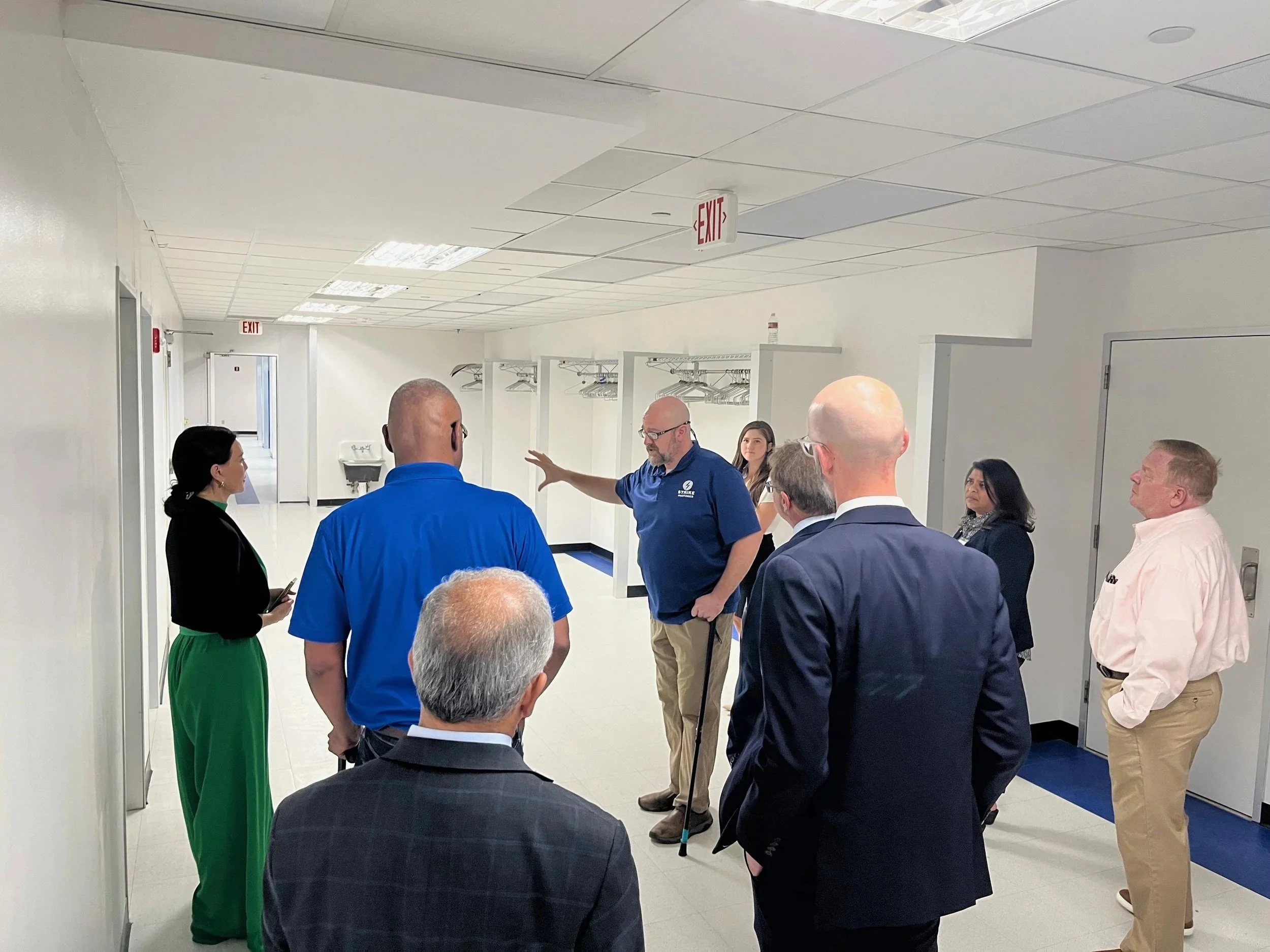 Group of people standing and talking in a hallway with white walls, tiled ceiling, and lockers in the background.