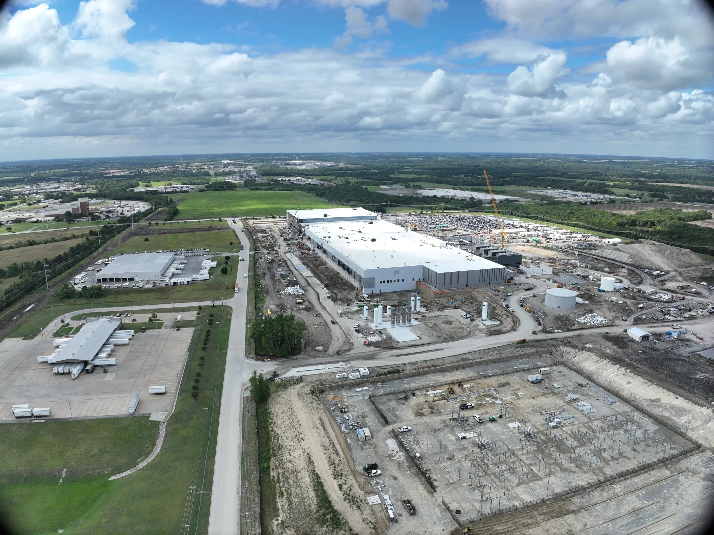 Aerial view of a large construction site with a white warehouse building, construction cranes, ongoing work, and surrounding infrastructure, in a green and rural landscape with roads, fields, and trees.