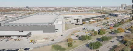 Aerial view of a large industrial or warehouse complex with surrounding roads and parking lot, cityscape in the background.