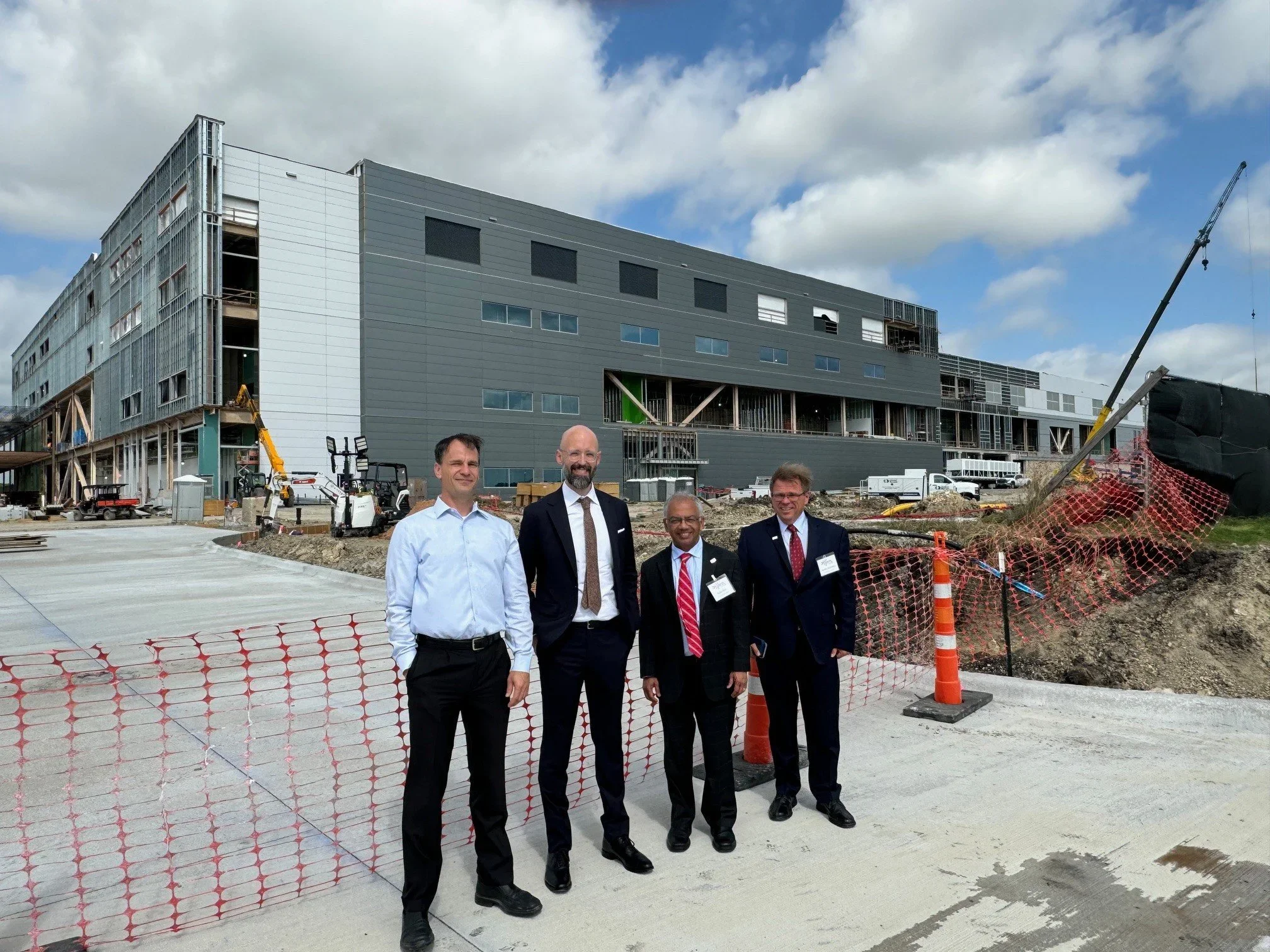 Four men in formal attire standing at a construction site with a large, modern building under construction in the background. The site is fenced with orange safety netting and cones, and construction equipment and vehicles are visible.