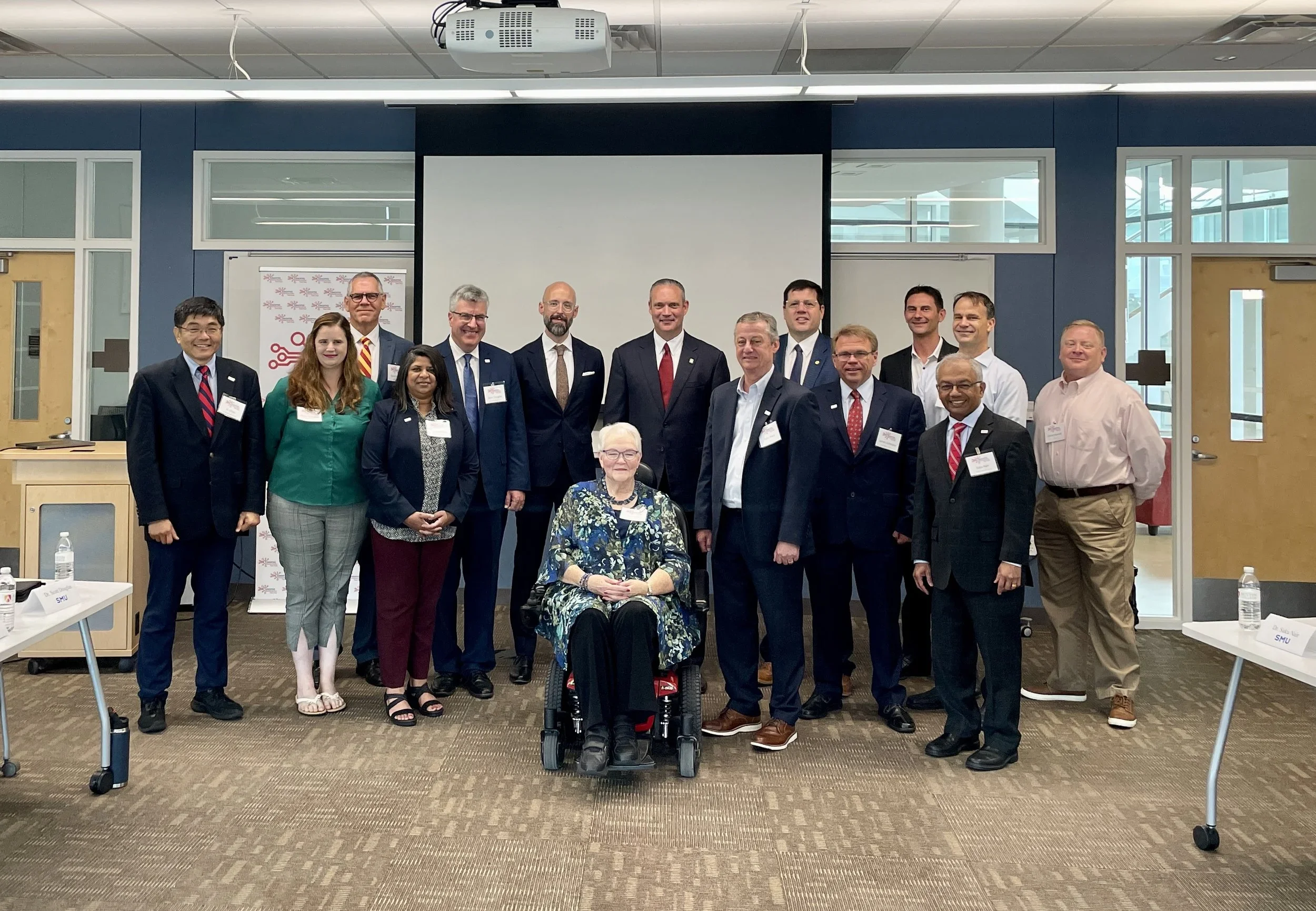 Group of diverse professionals posing for a photo in a conference room, some wearing name tags, with a large screen and a banner in the background.