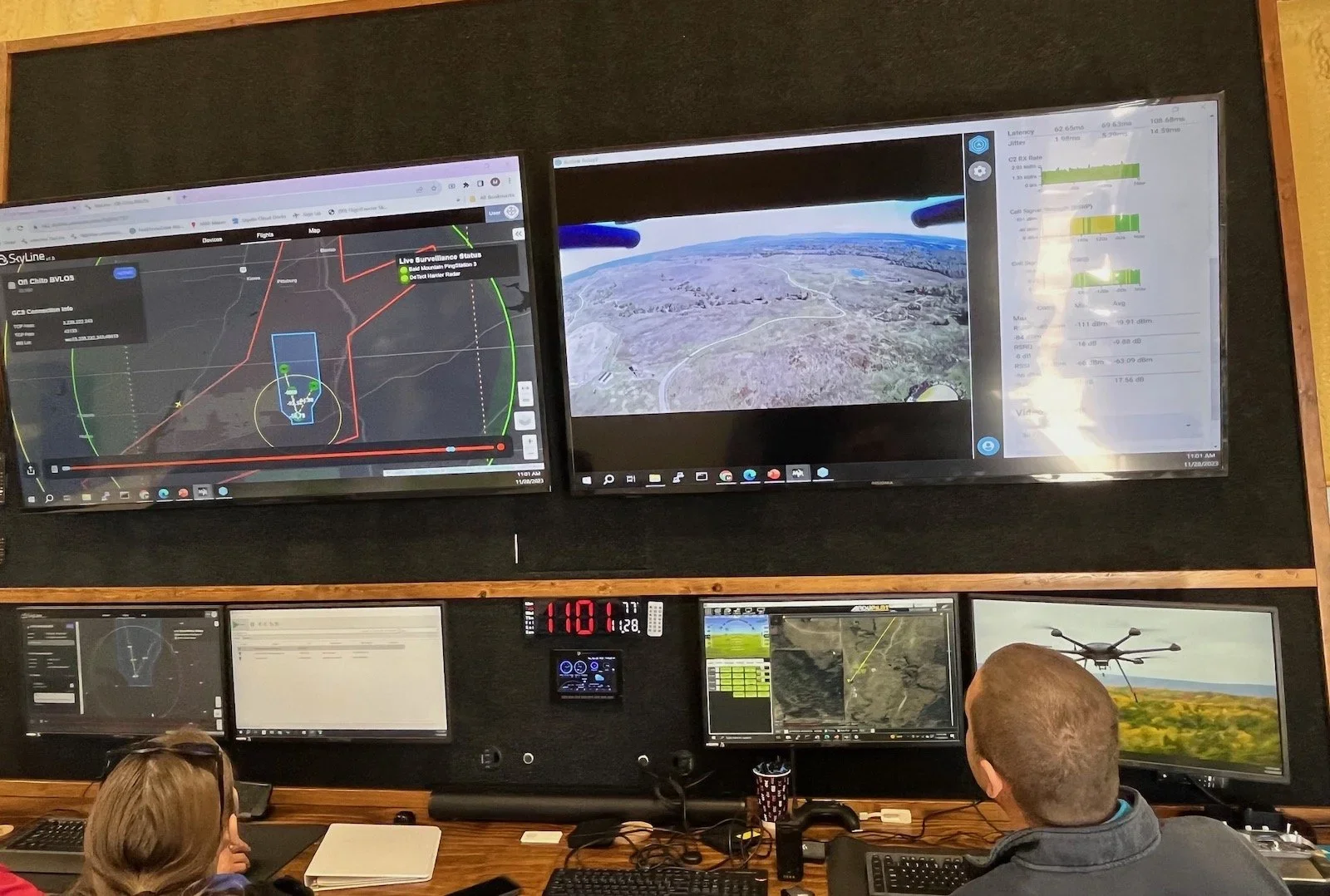 Two people in a control room looking at multiple monitors displaying drone flight paths, real-time camera footage, and telemetry data.