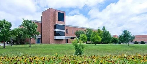 A large brick building with a prominent tower, surrounded by a green lawn and trees, under a partly cloudy sky.
