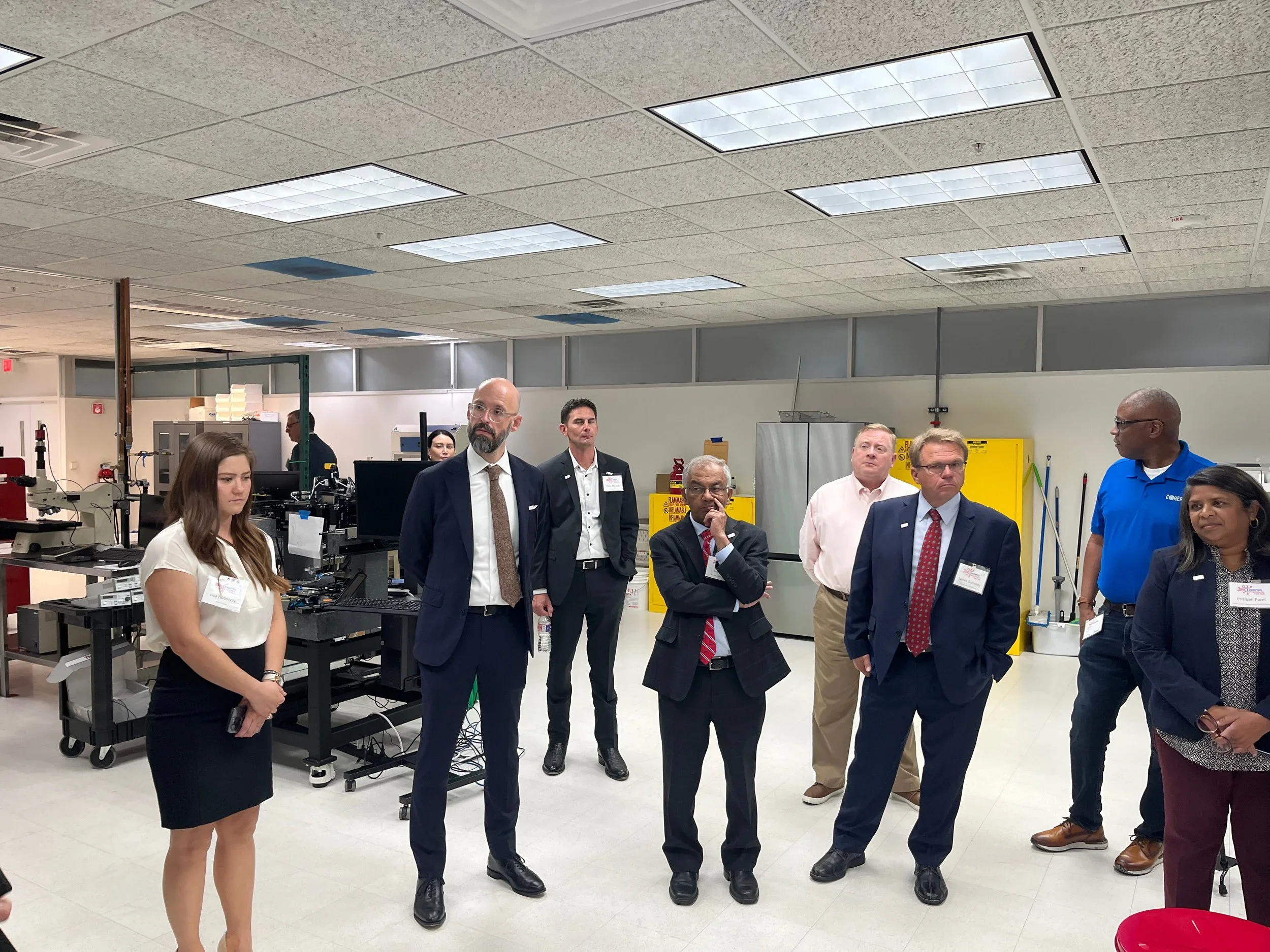 Group of professionals in a laboratory or workshop setting, standing and listening attentively, with equipment and cabinets in the background.