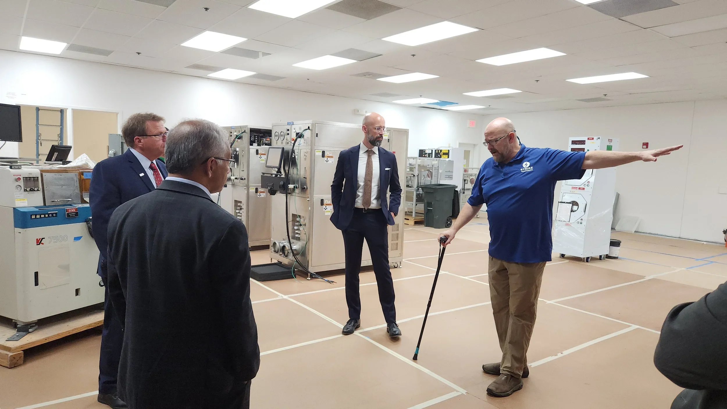 A group of four men in business attire and one man in a blue polo shirt with a logo, in a room with industrial equipment. One man is giving a presentation or explanation, gesturing with his arm extended.