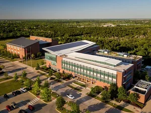 An aerial view of a large modern commercial building with glass windows, surrounded by parking lots filled with cars, trees, and open green space, with a forest in the background.