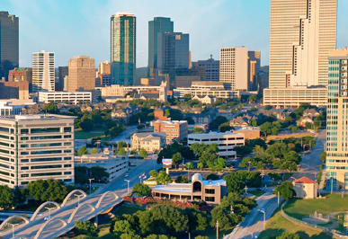 A city skyline with tall modern skyscrapers and lower buildings, with green trees and a clear sky.
