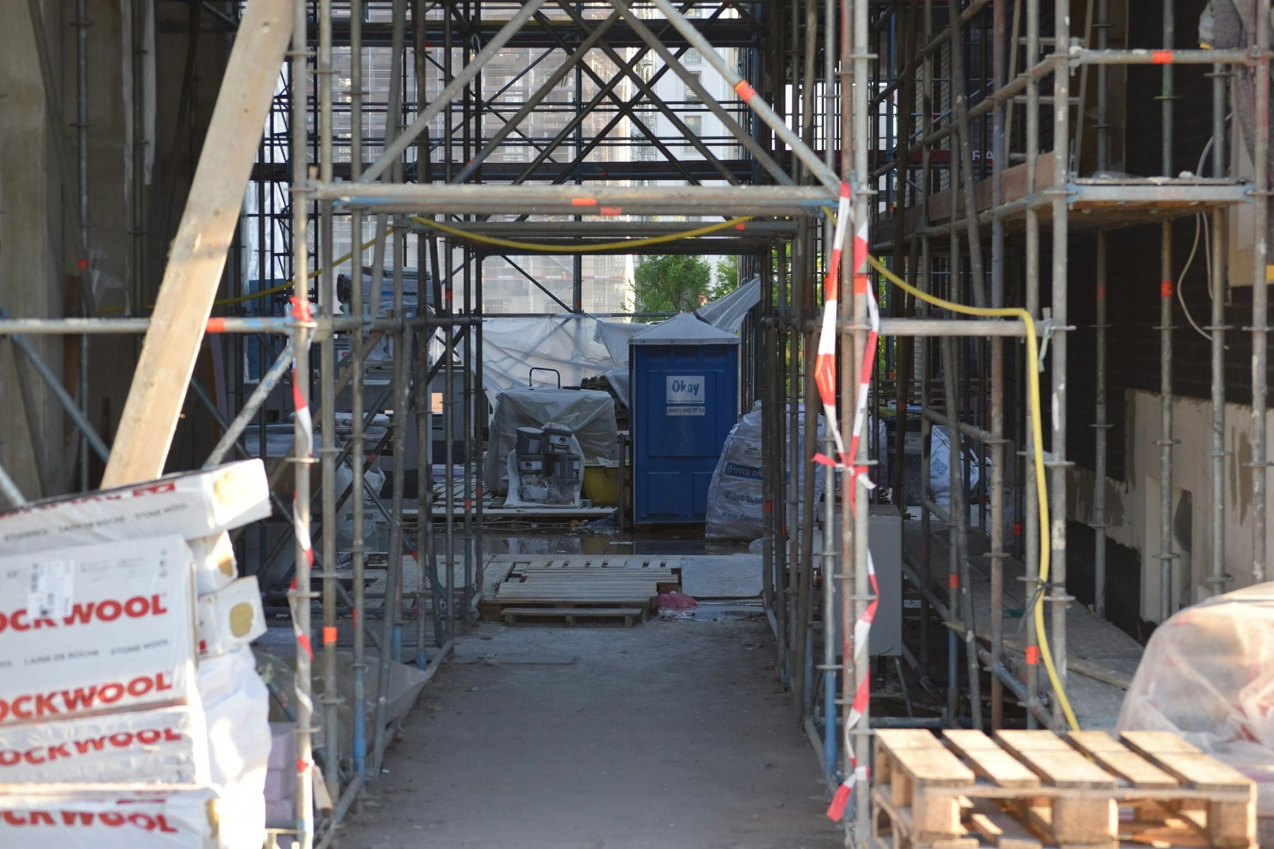 Construction site with metal scaffolding, bags of insulation, and building materials arranged around the area with a portable toilet showing between the opening of the scaffolding.