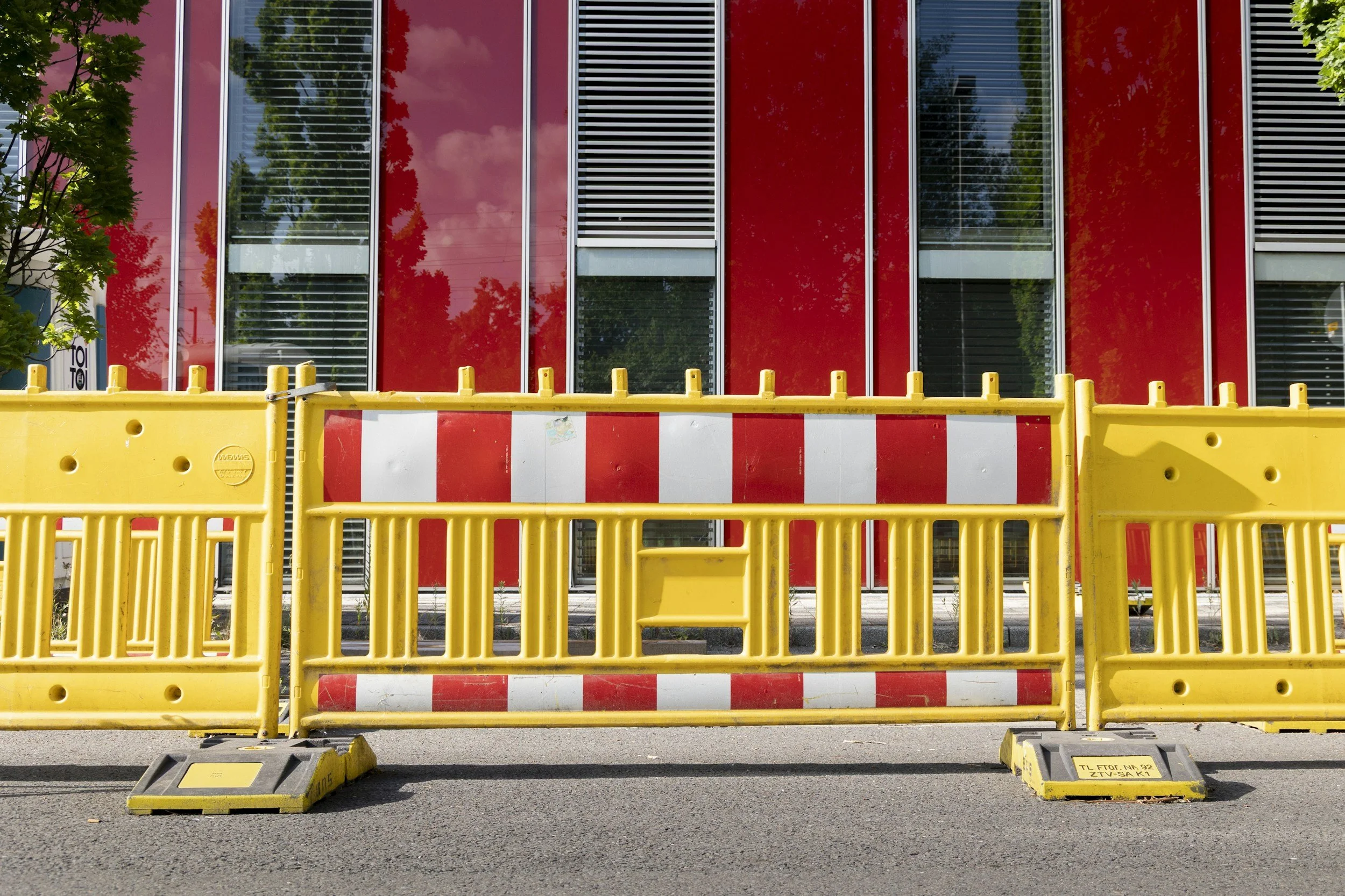A building with red and glass exterior walls, behind a yellow construction barrier on a paved street.