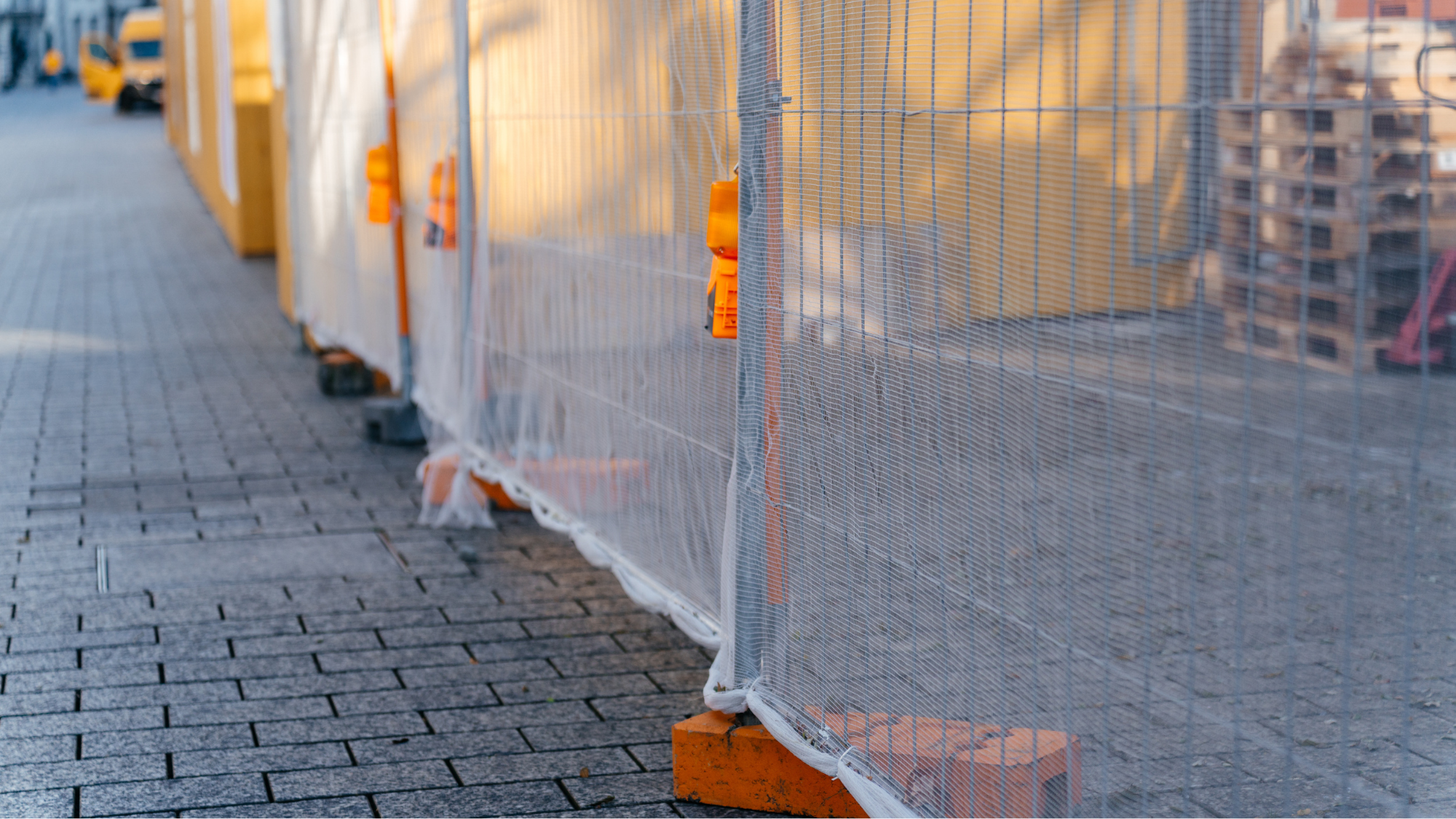 Construction site barrier made of metal mesh with orange bricks and yellow fencing, situated on a paved sidewalk.