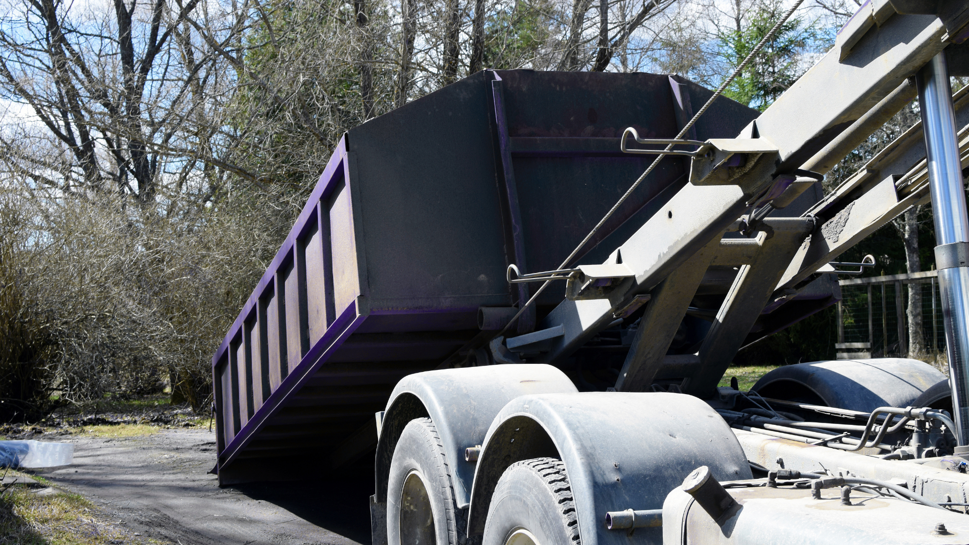 A dump truck with a raised bed, partially seen in a rural or construction area with trees and a fence in the background.