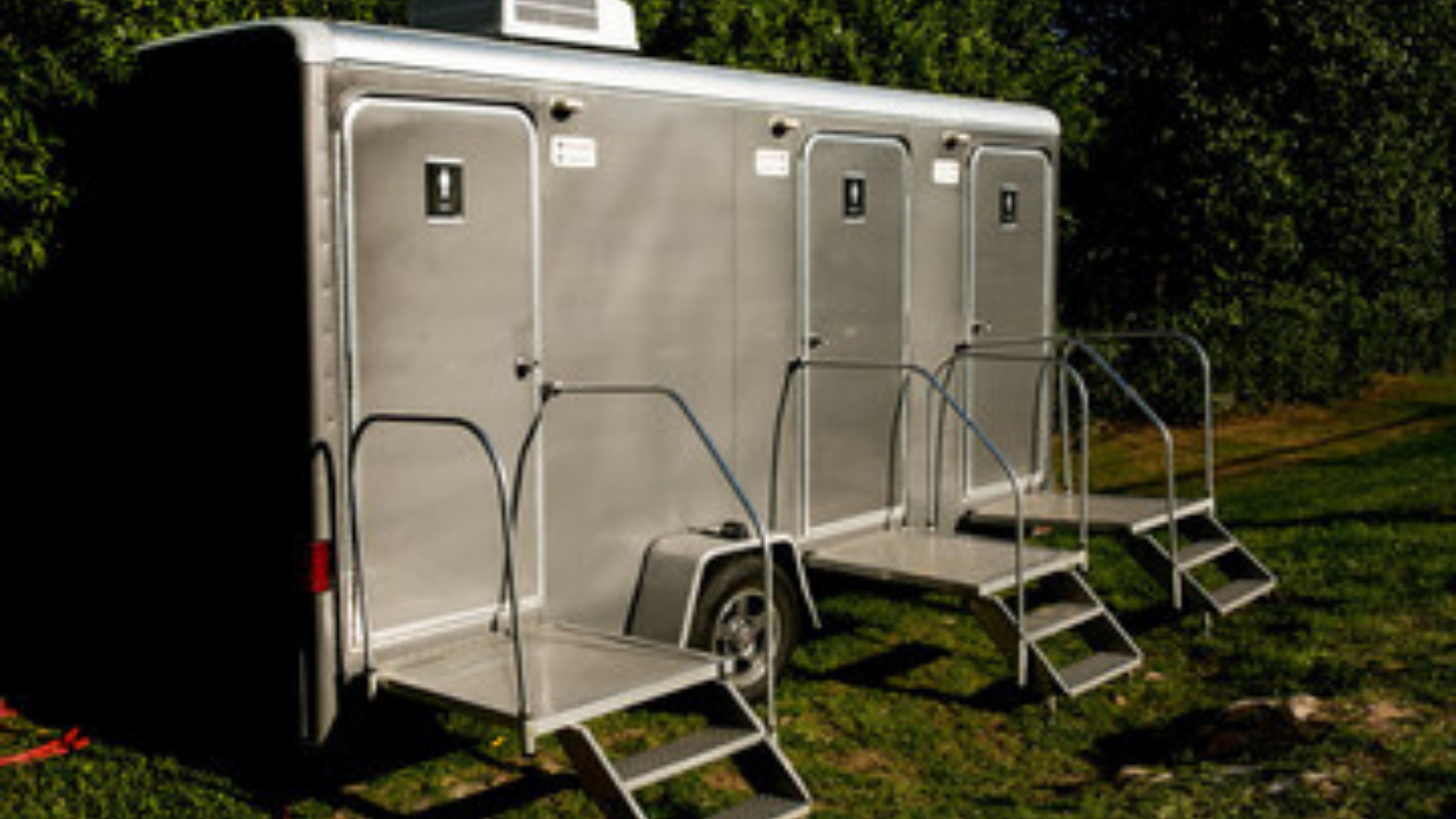 Steel luxury portable toilet trailer with three stairs leading up to three separate doors, parked on grass with bushes in the background.
