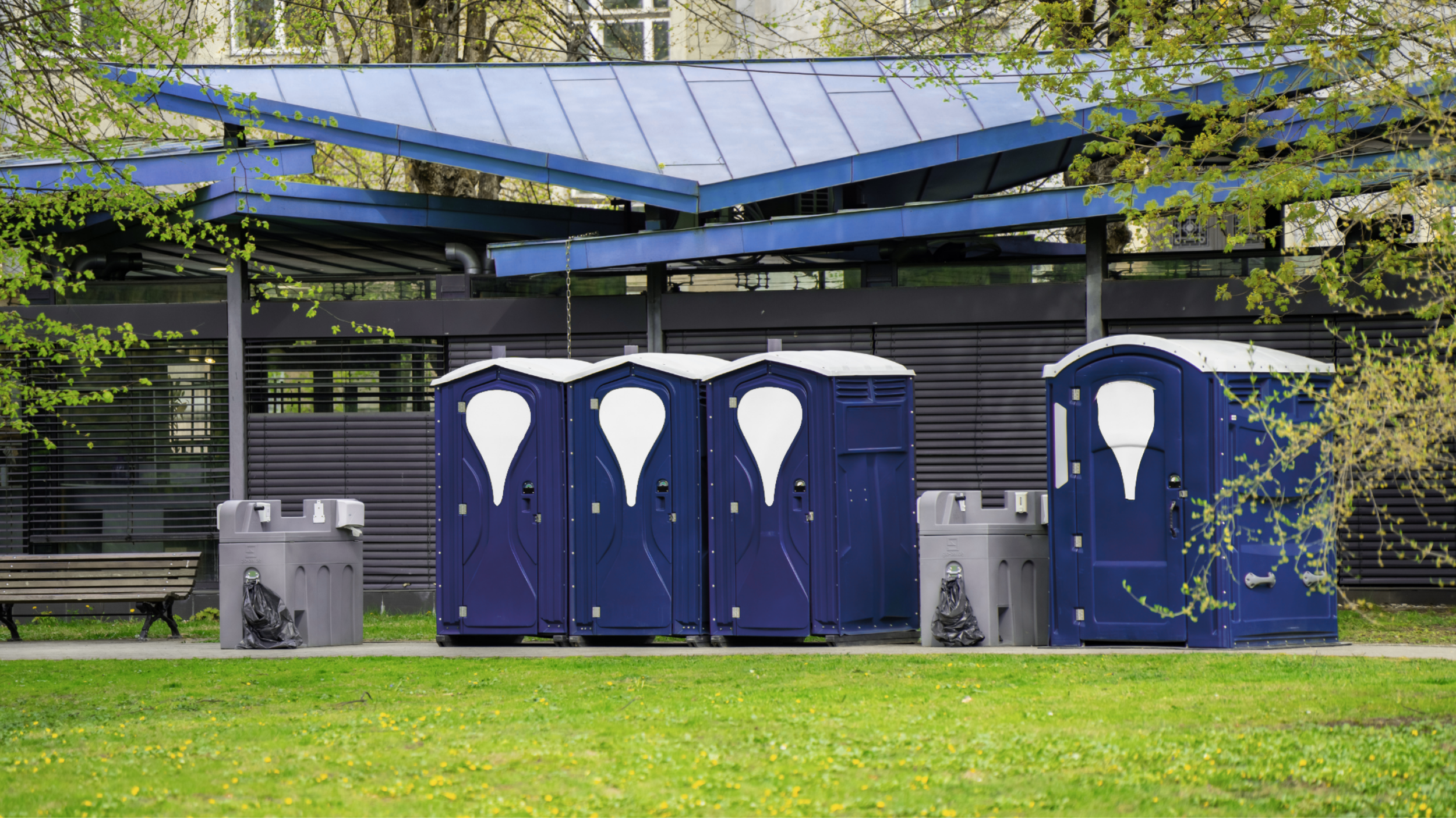Four portable toilets and two hand sanitizing stations in a park with blue portable toilets, grey hand sanitizing stations, trash cans, a park bench, and a modern building with a blue roof and trees in the background.