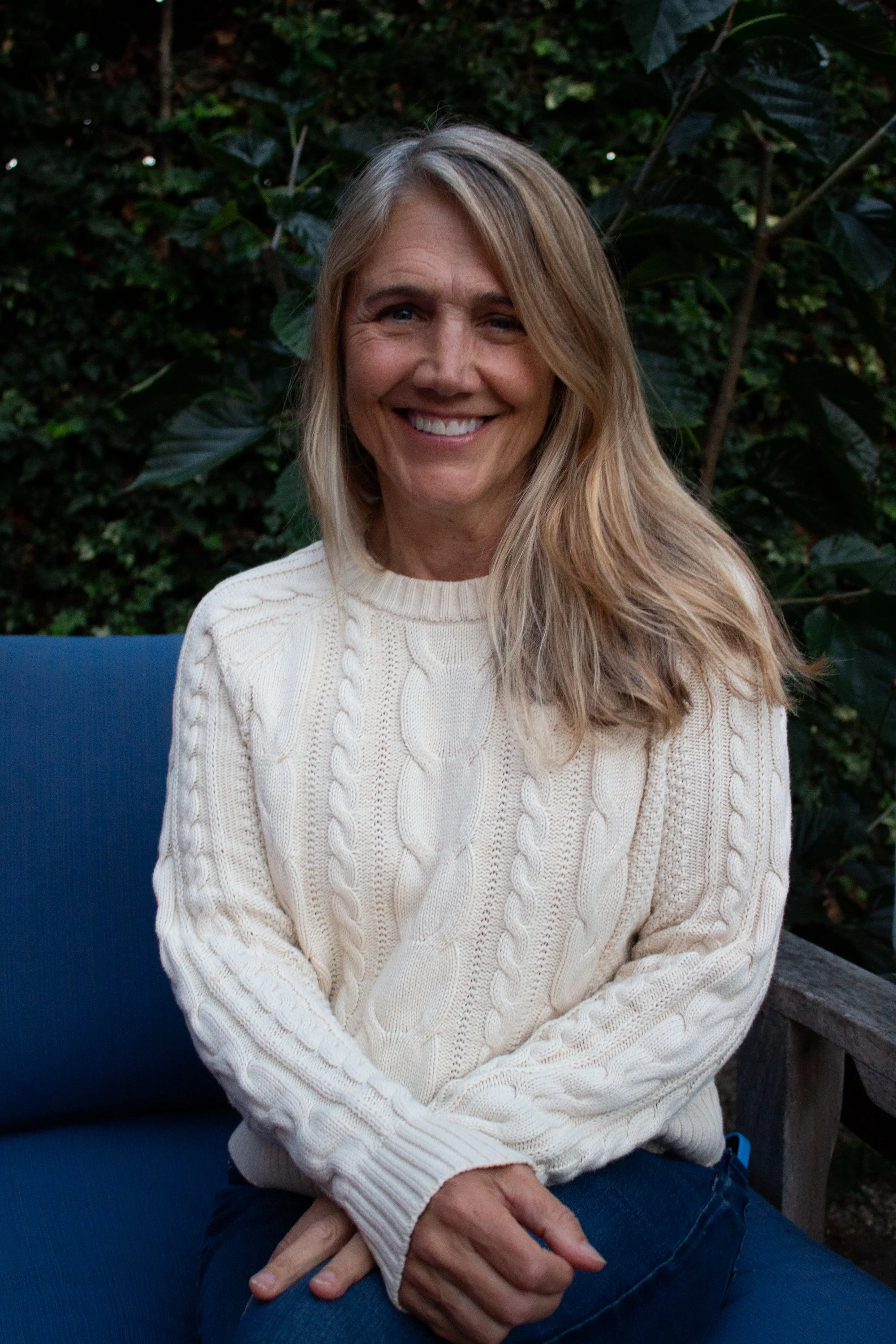 A smiling woman with blonde hair wearing a white cable-knit sweater, sitting outdoors in front of green foliage.