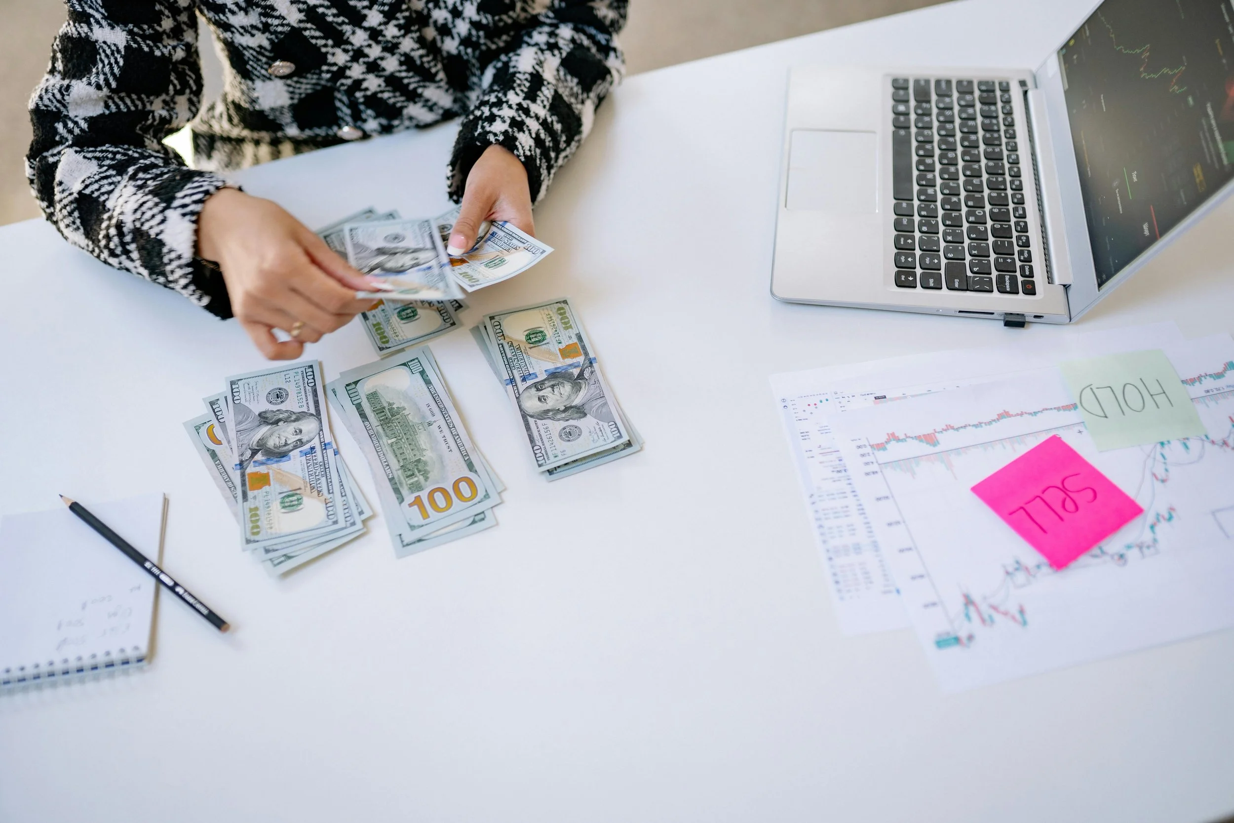 Person counting stacks of 100-dollar bills at a desk with a laptop, stock charts, and notes.