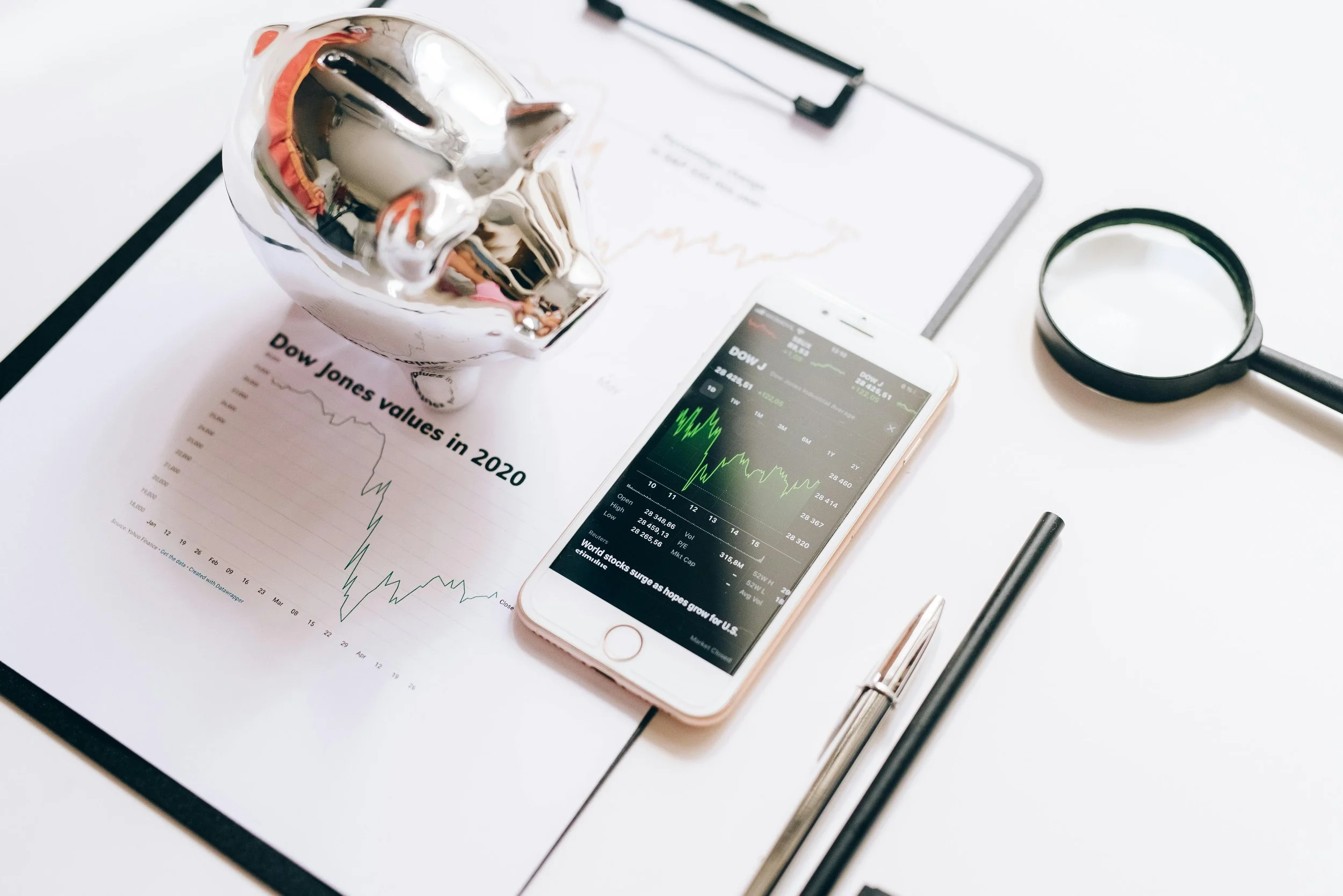 Work desk with a chart titled 'Dow Jones values in 2020', a smartphone displaying stock market data, a magnifying glass, a black pen, a silver pen, and a reflective silver piggy bank.