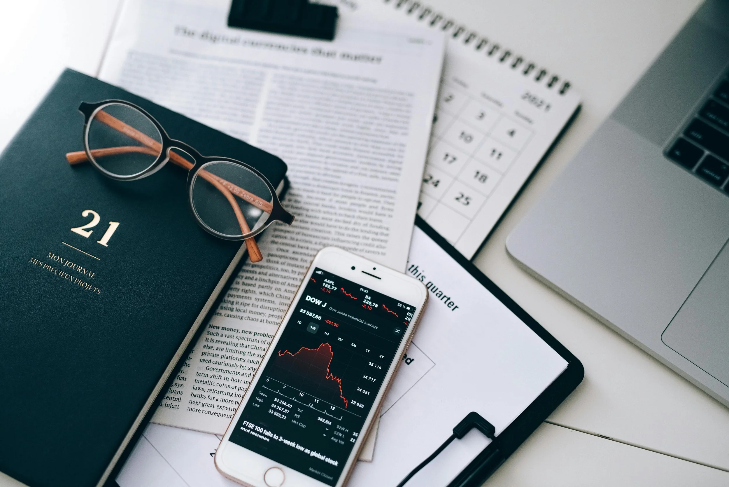A workspace with a closed notebook, a pair of eyeglasses, a smartphone displaying stock market data, a calendar, a printed newspaper, a laptop, and a clipboard on a white desk.