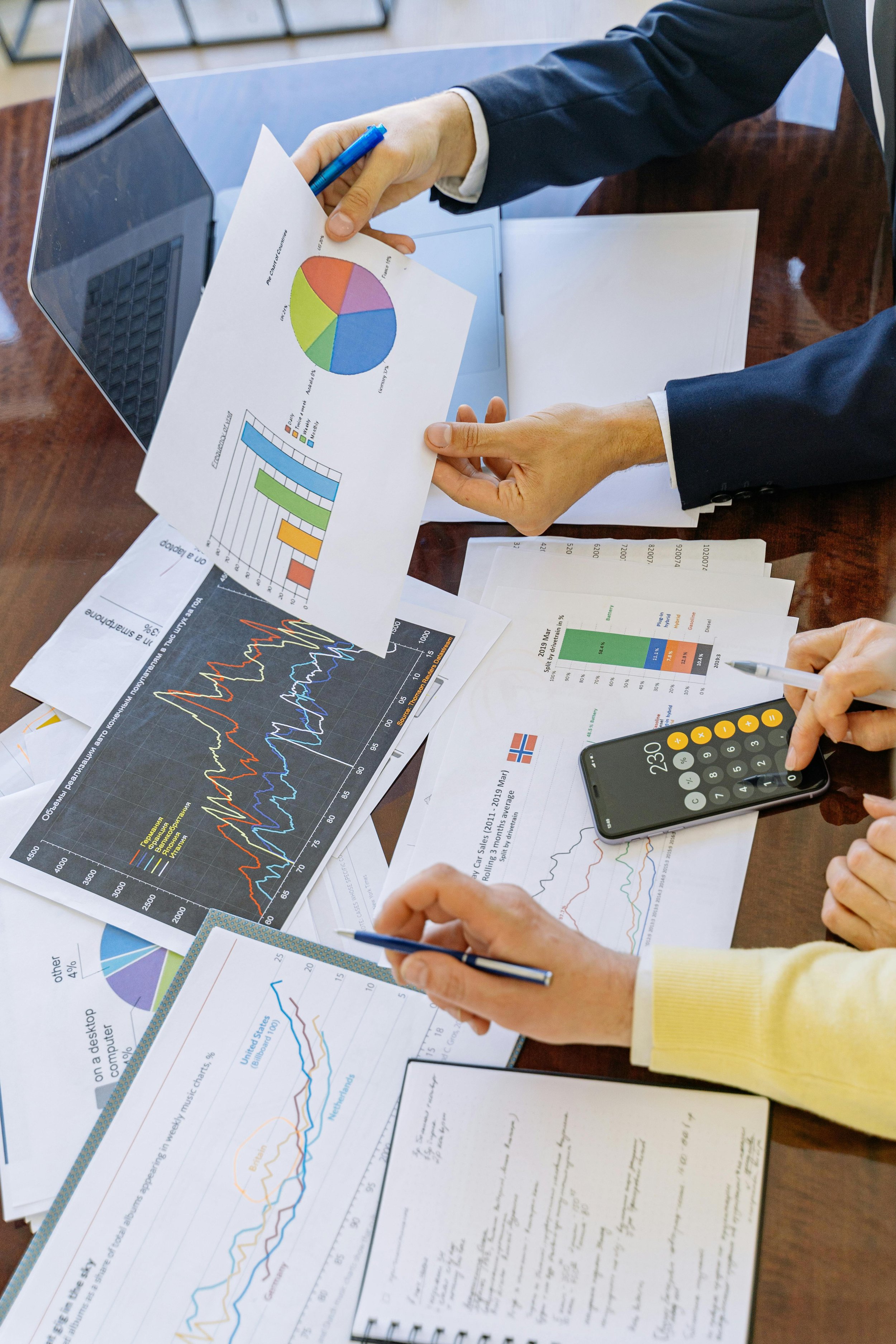 People working on financial and statistical reports with printed charts, graphs, a laptop, and a calculator on a wooden table.