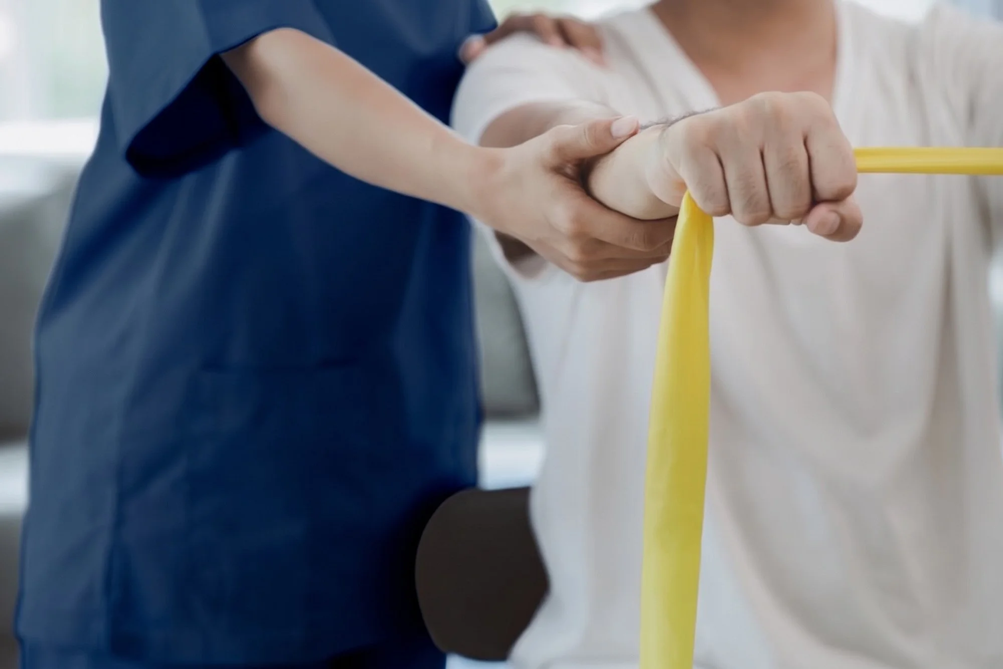 Physiotherapist in navy scrubs supporting a patient's shoulder and wrist as they stretch a yellow resistance band during a rehabilitation exercise.