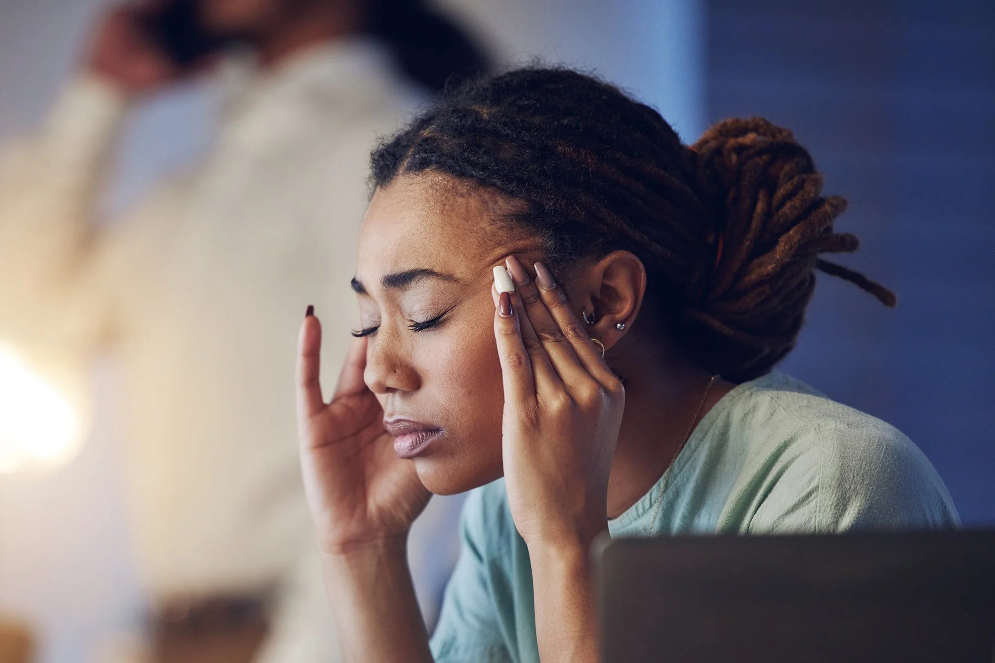 Woman at a laptop pressing her fingers to her temple with her eyes closed, showing signs of headache pain.