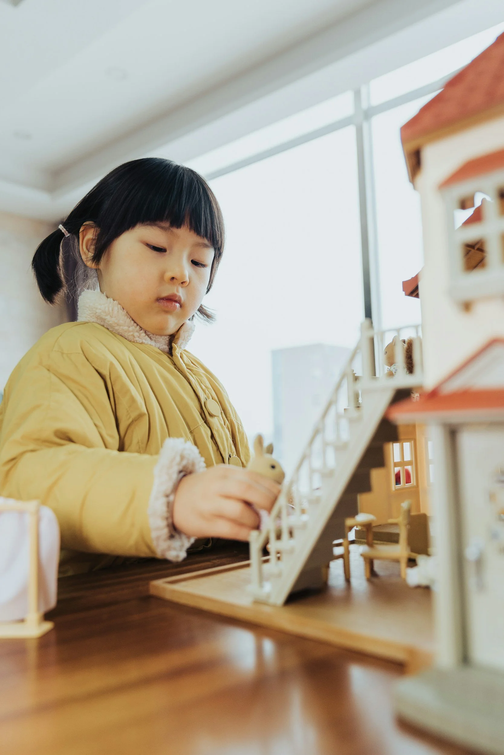 A young girl with black hair in pigtails wearing a yellow jacket playing with a wooden toy rabbit at a dollhouse indoors near a large window.