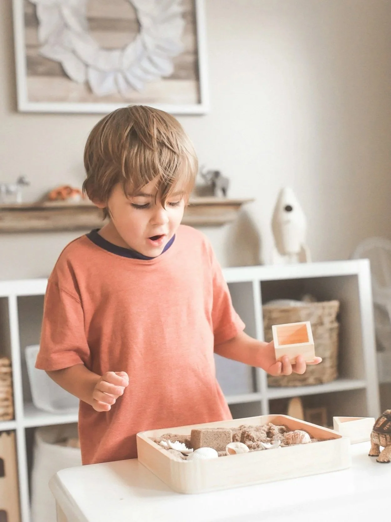 A young boy with light brown hair wearing an orange t-shirt standing at a table with a wooden tray filled with sand, shells, and small toys, looking surprised or excited.